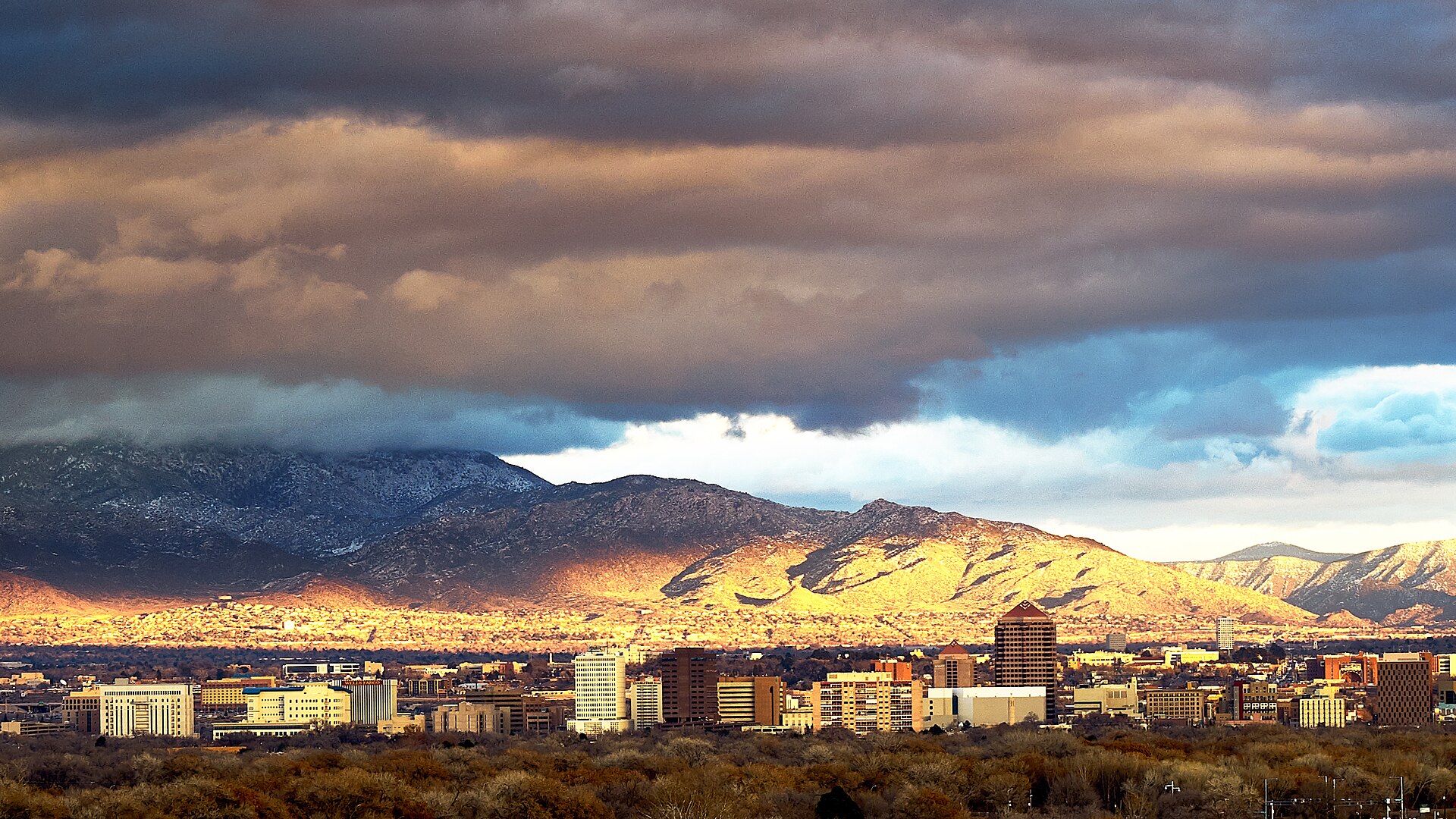 Downtown Albuquerque's skyline beneath the sunlit Sandia Mountains under dramatic clouds.