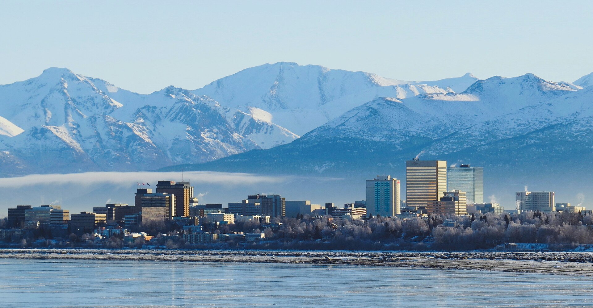 Downtown Anchorage's towers beneath the snow-covered Chugach Mountains, seen across icy water on a clear winter day.