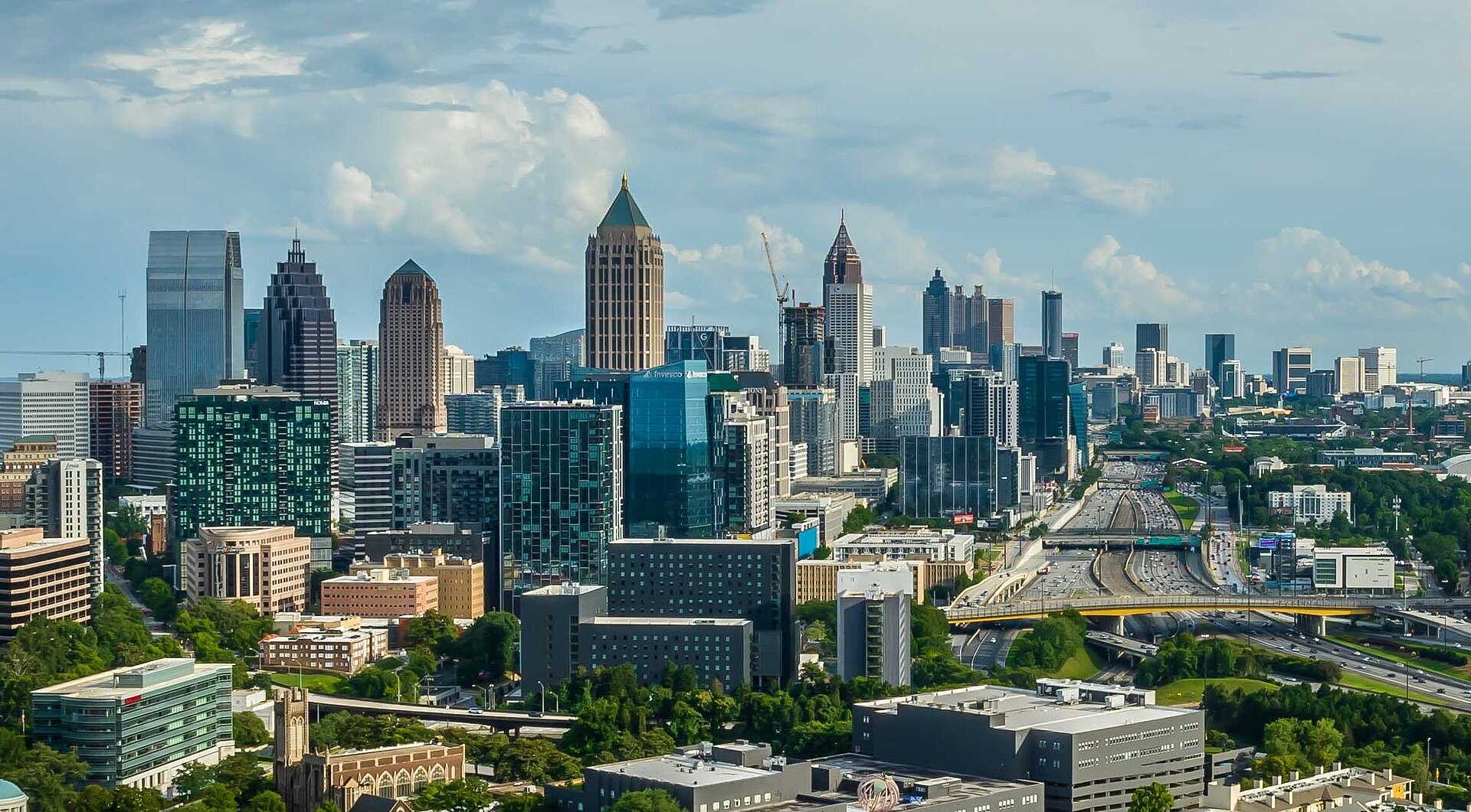 The skyline of Midtown and Downtown Atlanta rising above trees and the Downtown Connector under a partly cloudy sky.
