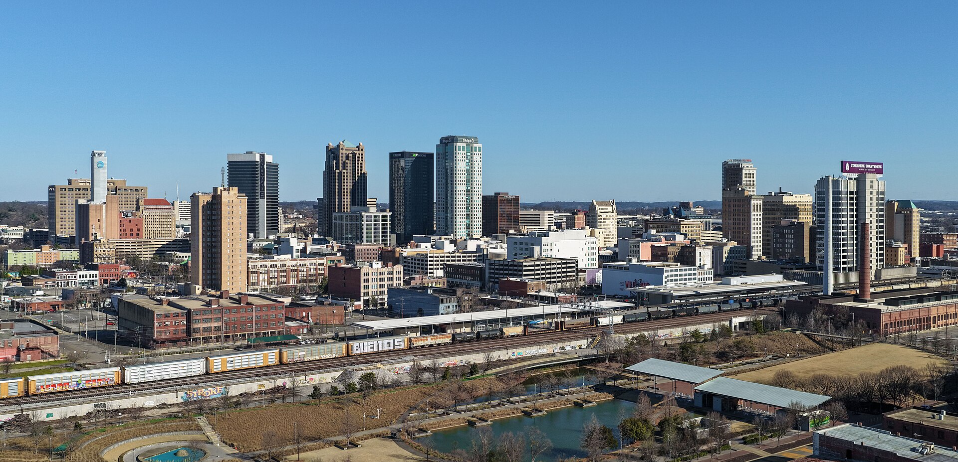 Downtown Birmingham skyline under a clear blue sky, with Railroad Park and the rail line in the foreground.