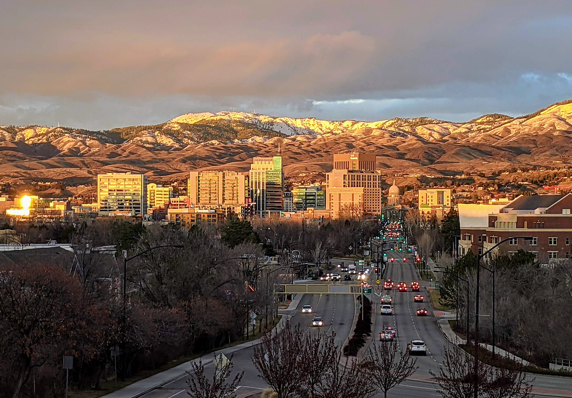 Capitol Boulevard leading into downtown Boise at golden hour, with snow-dusted foothills glowing behind the towers.