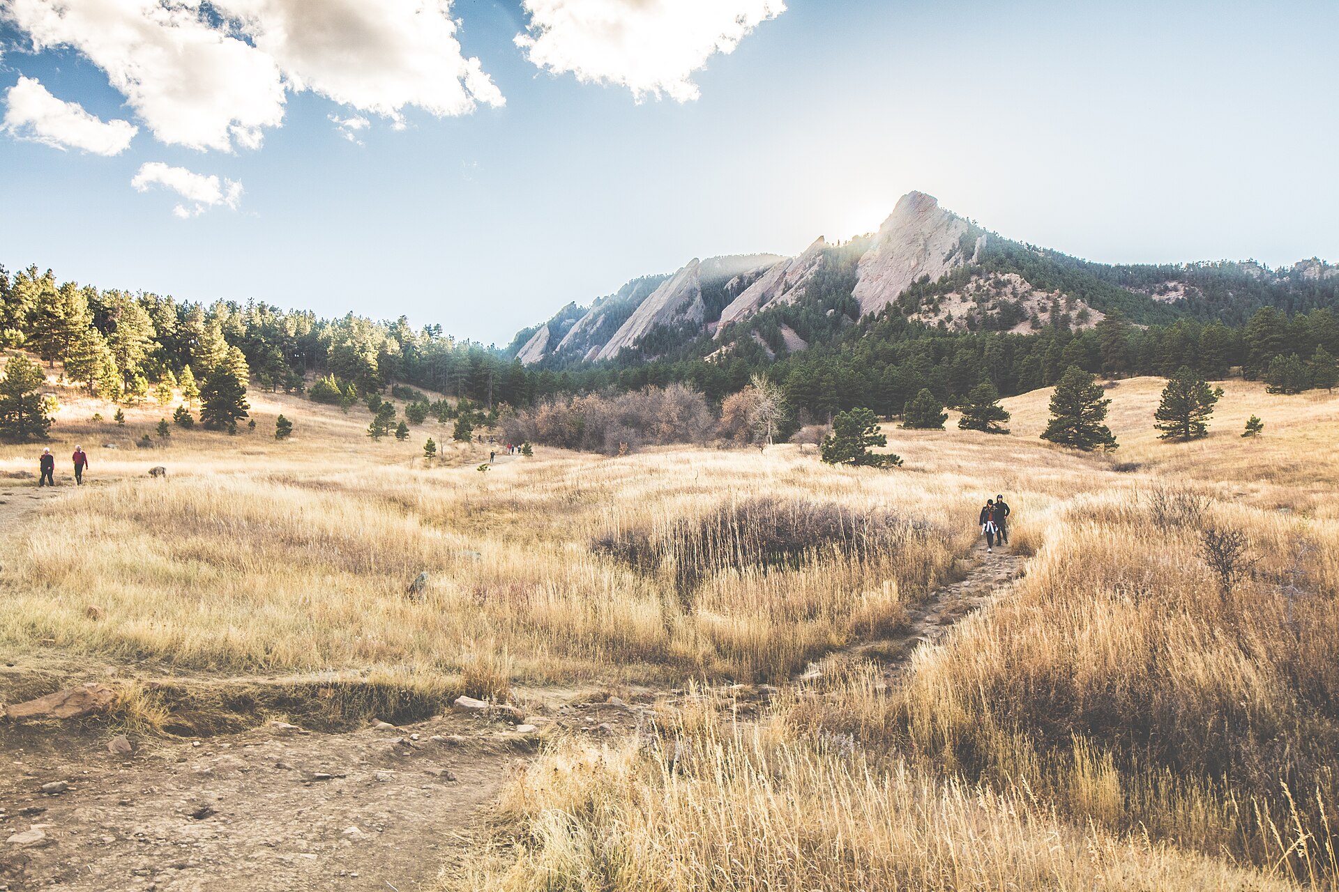 Hikers cross a golden grassy meadow below the Flatirons in Boulder on a sunny day.