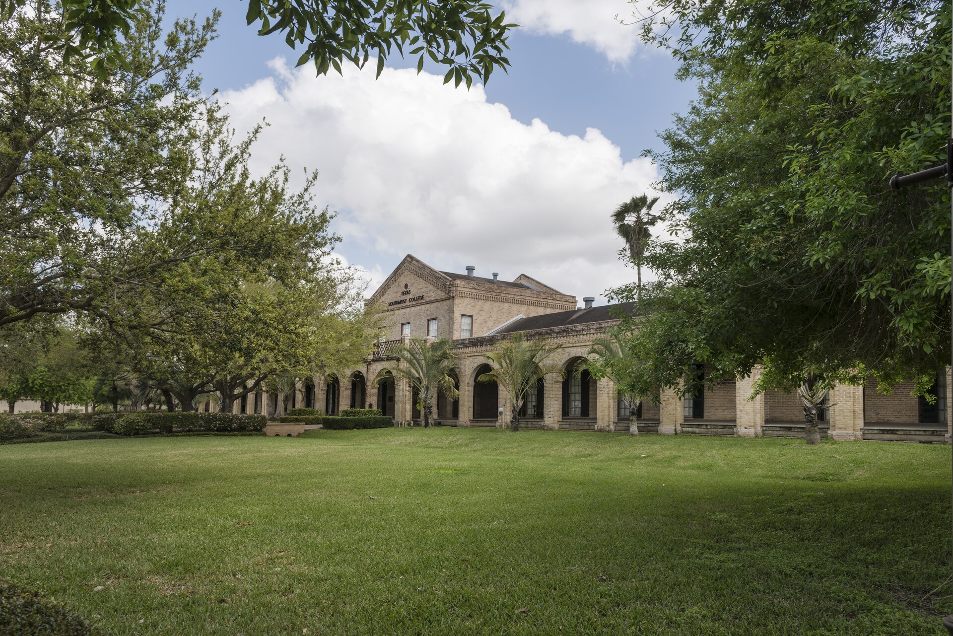Arcaded brick buildings of historic Fort Brown, now a university campus, behind a wide lawn, trees and palms in Brownsville.