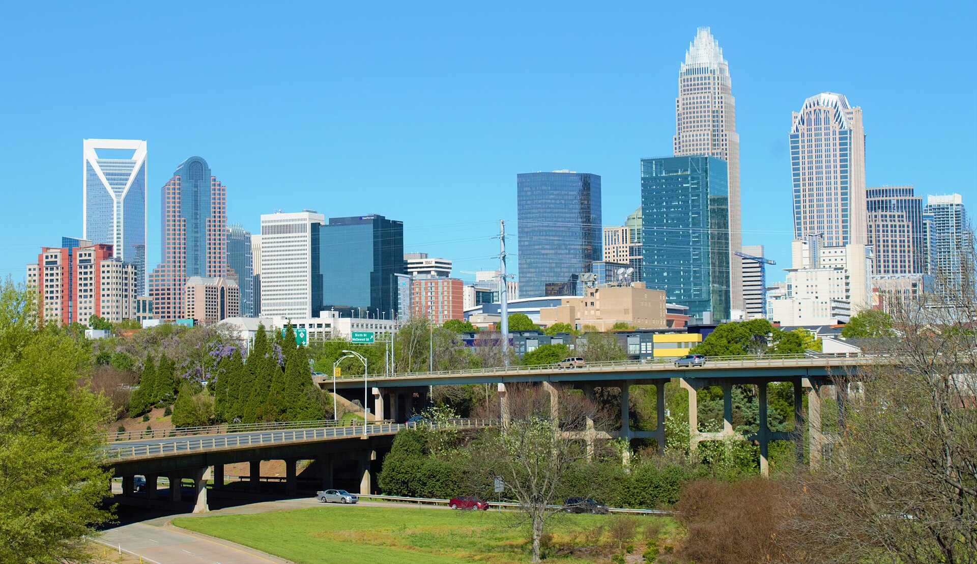 Uptown Charlotte's glass towers rising above spring trees and freeway overpasses under a clear blue sky.