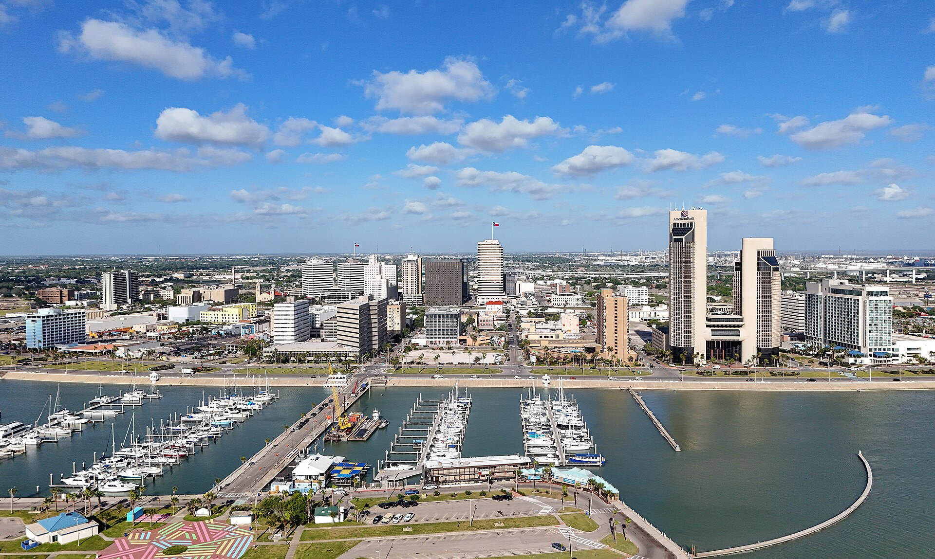 Marinas along the Corpus Christi bayfront below the downtown skyline under a blue sky with scattered clouds.