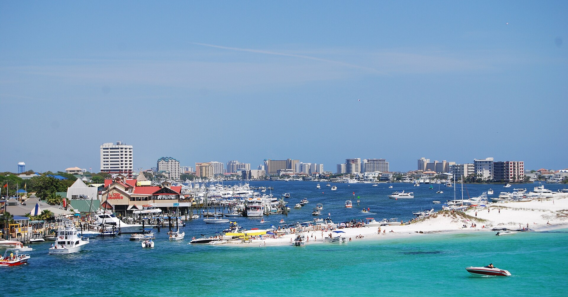 Boats, a white sandbar and emerald water at Destin Harbor, with beachfront towers beyond, on a sunny day.