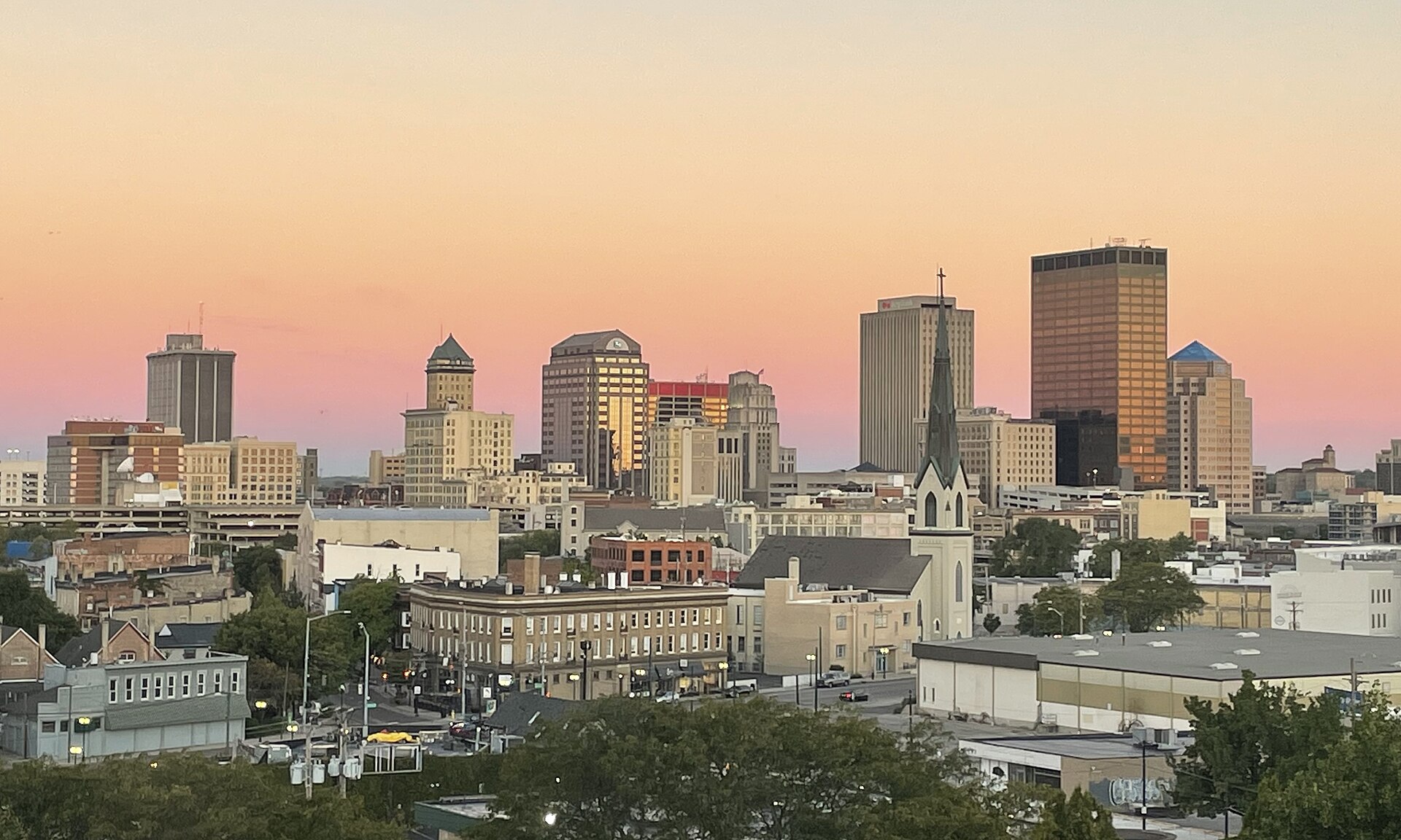 Downtown Dayton's skyline and a church steeple above older brick neighbourhoods under a pink and orange sunset sky.