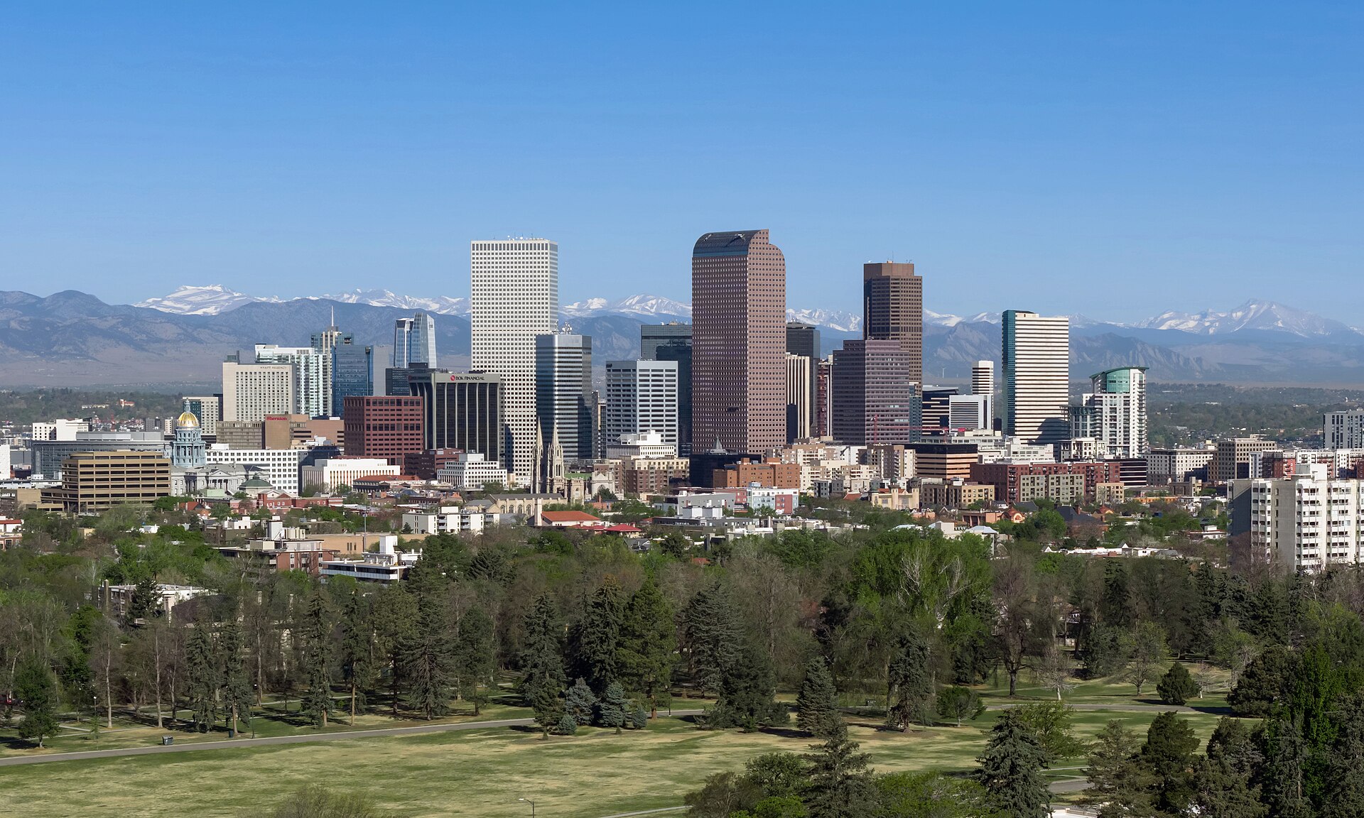 The downtown Denver skyline seen from City Park, with the snow-capped Rocky Mountains behind on a clear day.