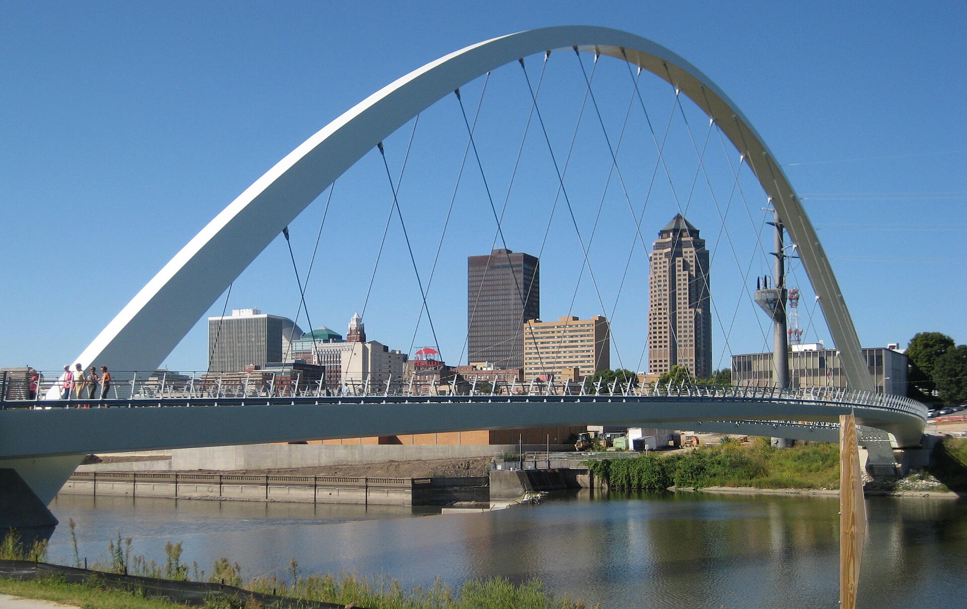 A white arched footbridge over the Des Moines River framing downtown Des Moines' towers under a clear blue sky.