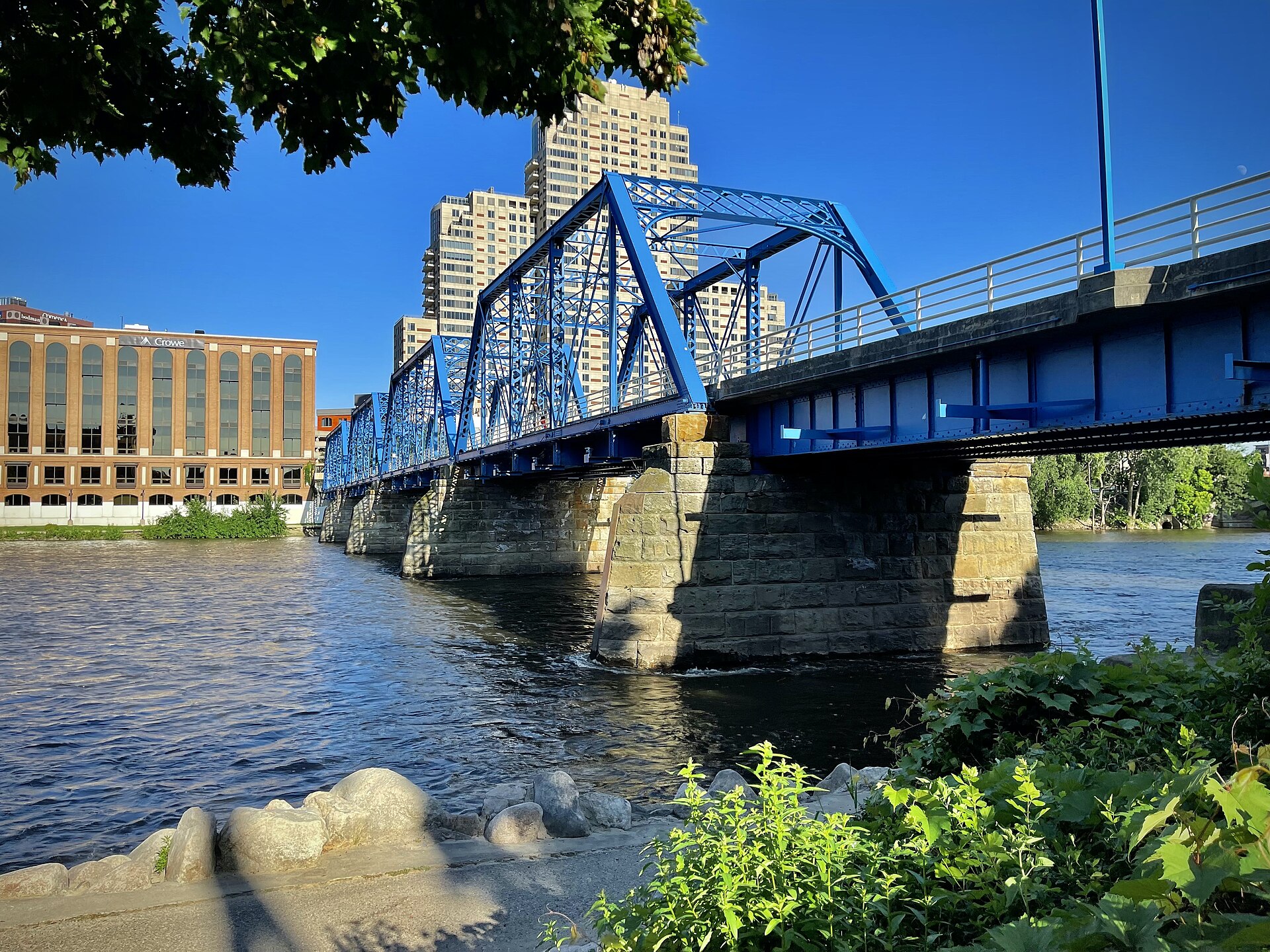 The Blue Bridge, a blue steel footbridge, crossing the Grand River toward downtown Grand Rapids under a clear sky.