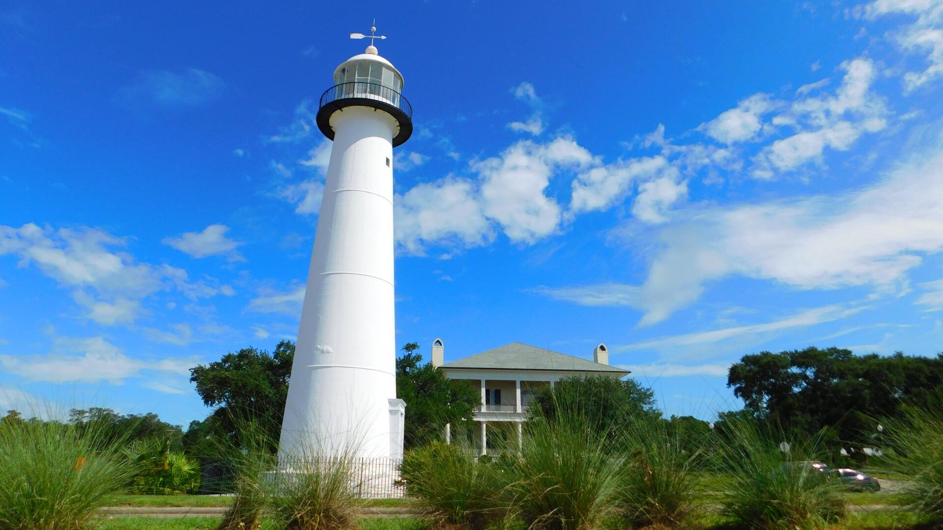 The white Biloxi Lighthouse beside the coast highway, with the Biloxi Visitors Center behind under a bright blue sky.