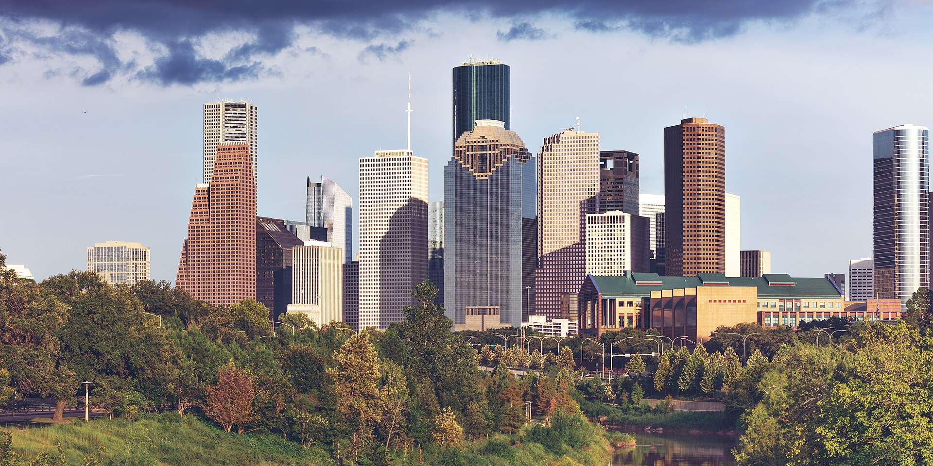 Downtown Houston skyscrapers rising above the green trees of Buffalo Bayou Park in late-afternoon sun.
