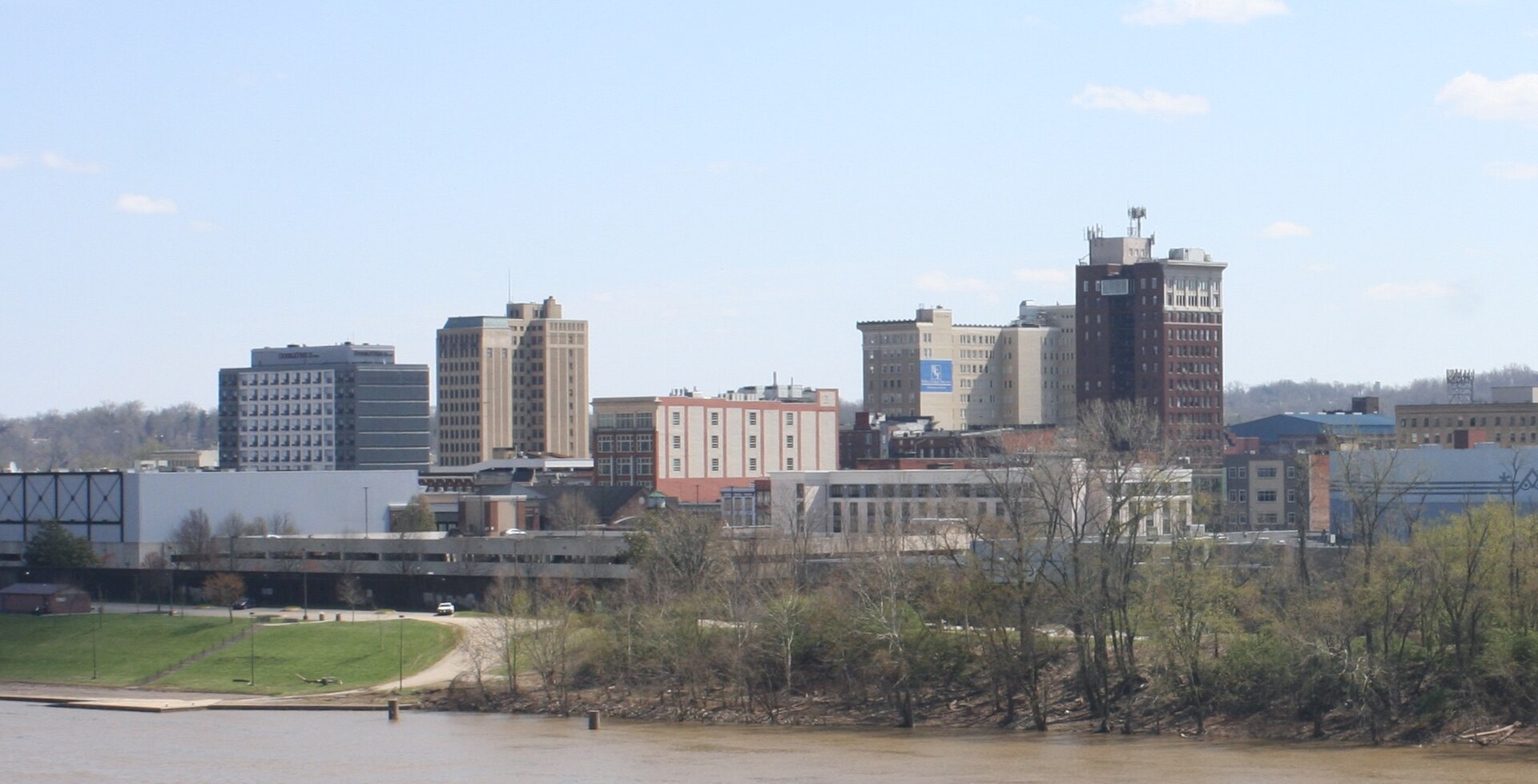 Downtown Huntington's buildings rise beyond the trees on the bank of the Ohio River under a blue sky.
