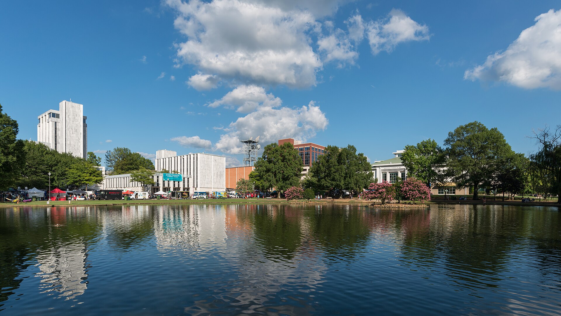 Downtown Huntsville buildings and trees reflected in the lagoon at Big Spring International Park under a blue summer sky.