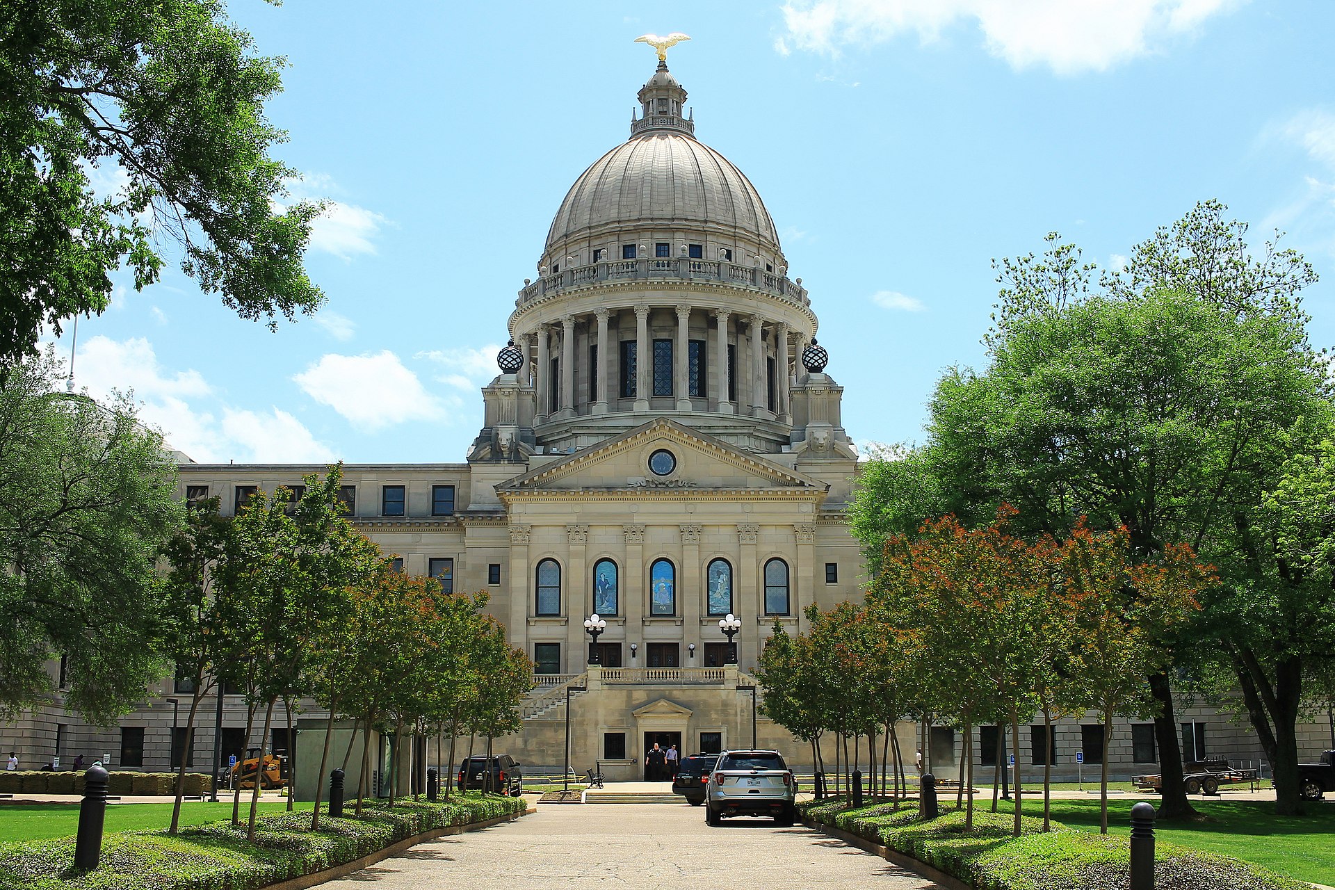 The domed Mississippi State Capitol in Jackson, framed by trees along its front walk on a sunny day.