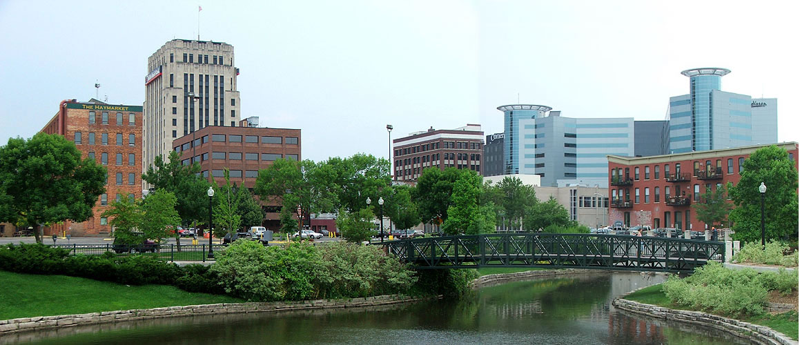 Downtown Kalamazoo's brick buildings and glass towers above a green footbridge over Arcadia Creek on a summer day.