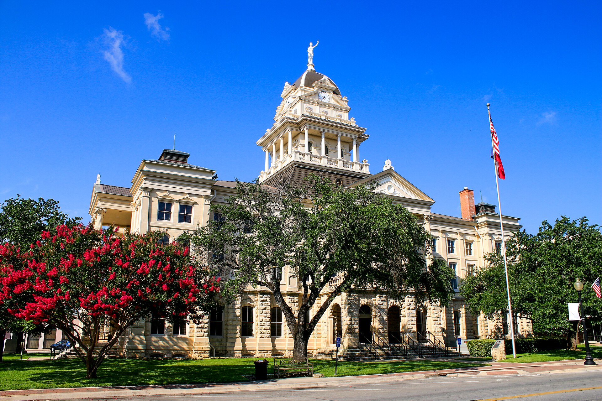 The 1884 Bell County Courthouse in Belton, with its domed clock tower, behind red crape myrtles under a deep blue sky.