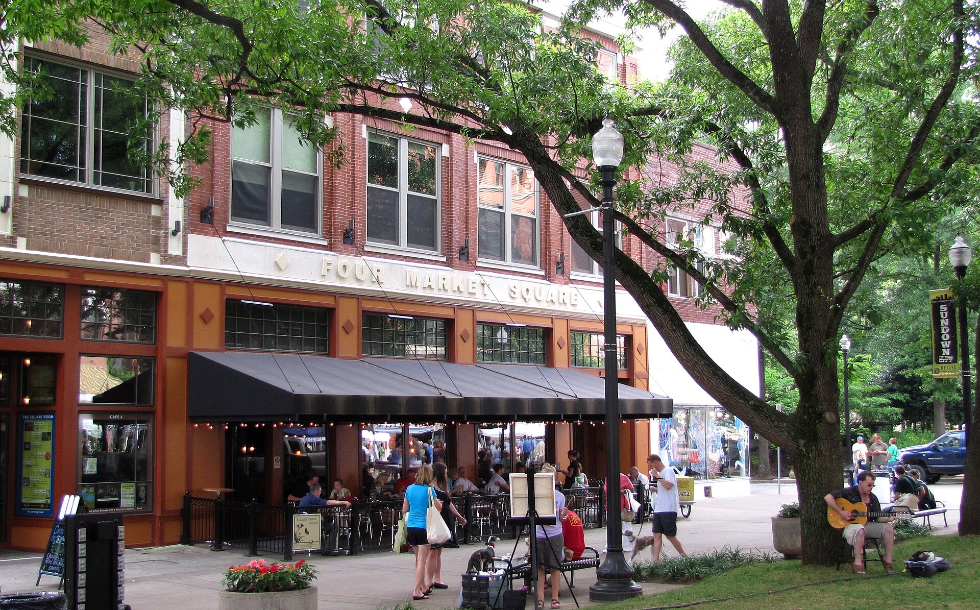 People strolling past brick buildings and cafe awnings under shade trees in Market Square, downtown Knoxville.