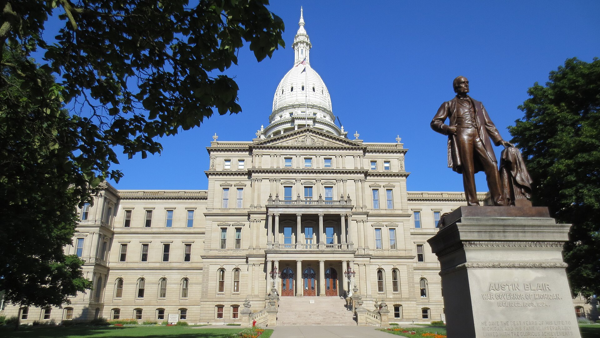 The domed Michigan State Capitol in Lansing under a clear blue sky, with the Austin Blair statue at right.