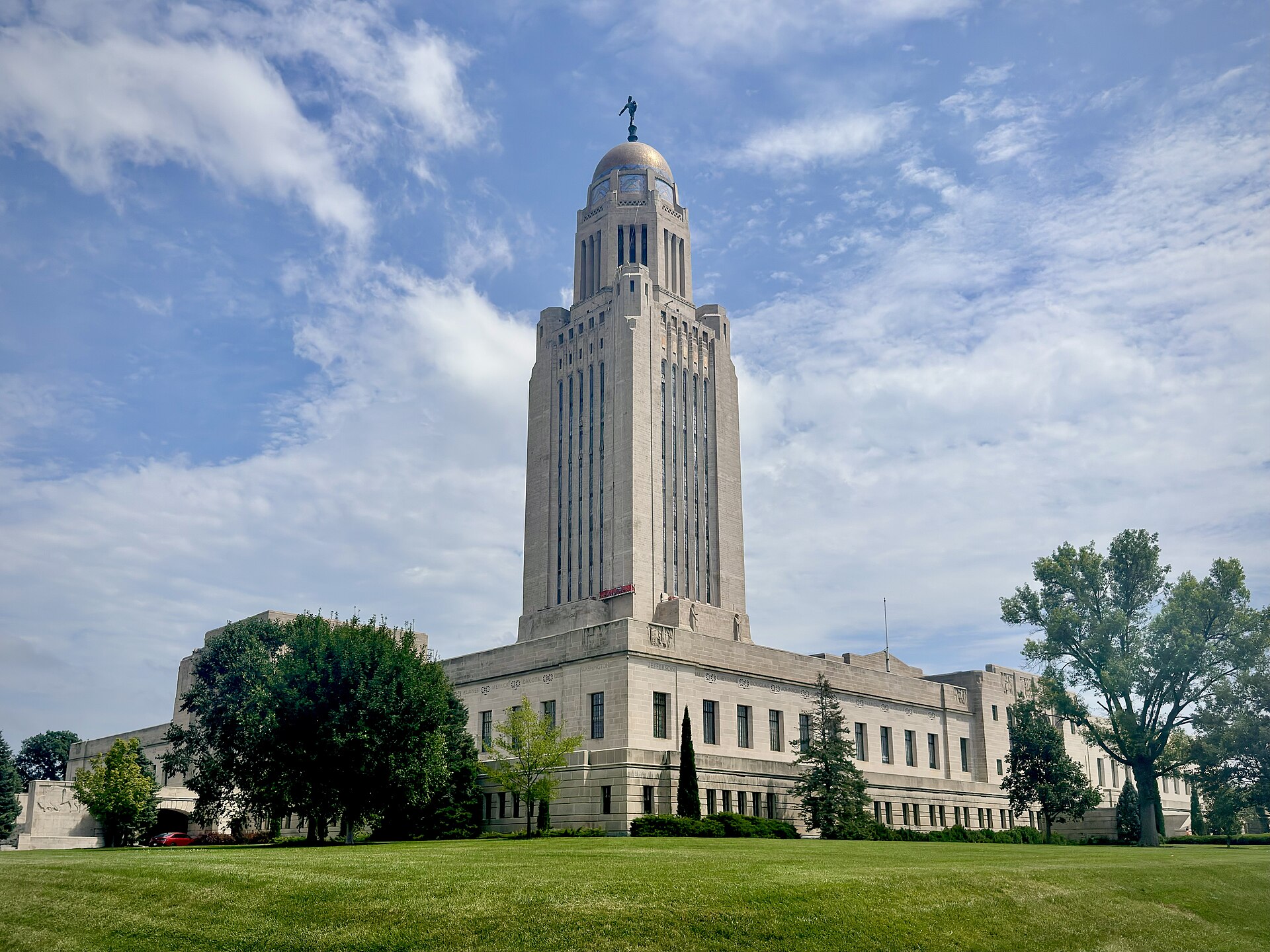 The Nebraska State Capitol's tall stone tower and domed top rise above trees and a green lawn under a blue, cloud-streaked sky.