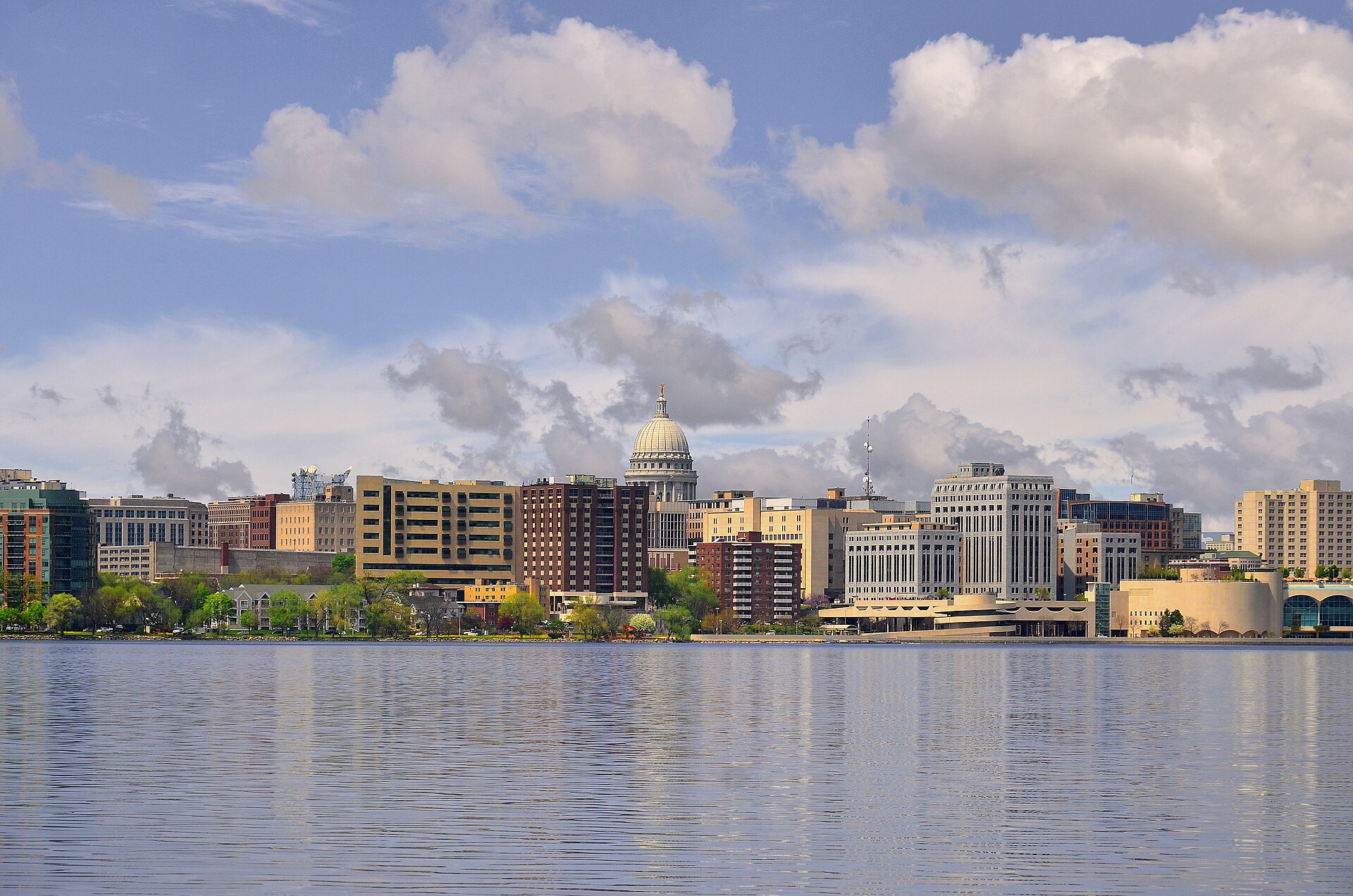Downtown Madison and the domed Wisconsin State Capitol seen across Lake Monona under a blue sky with puffy clouds.