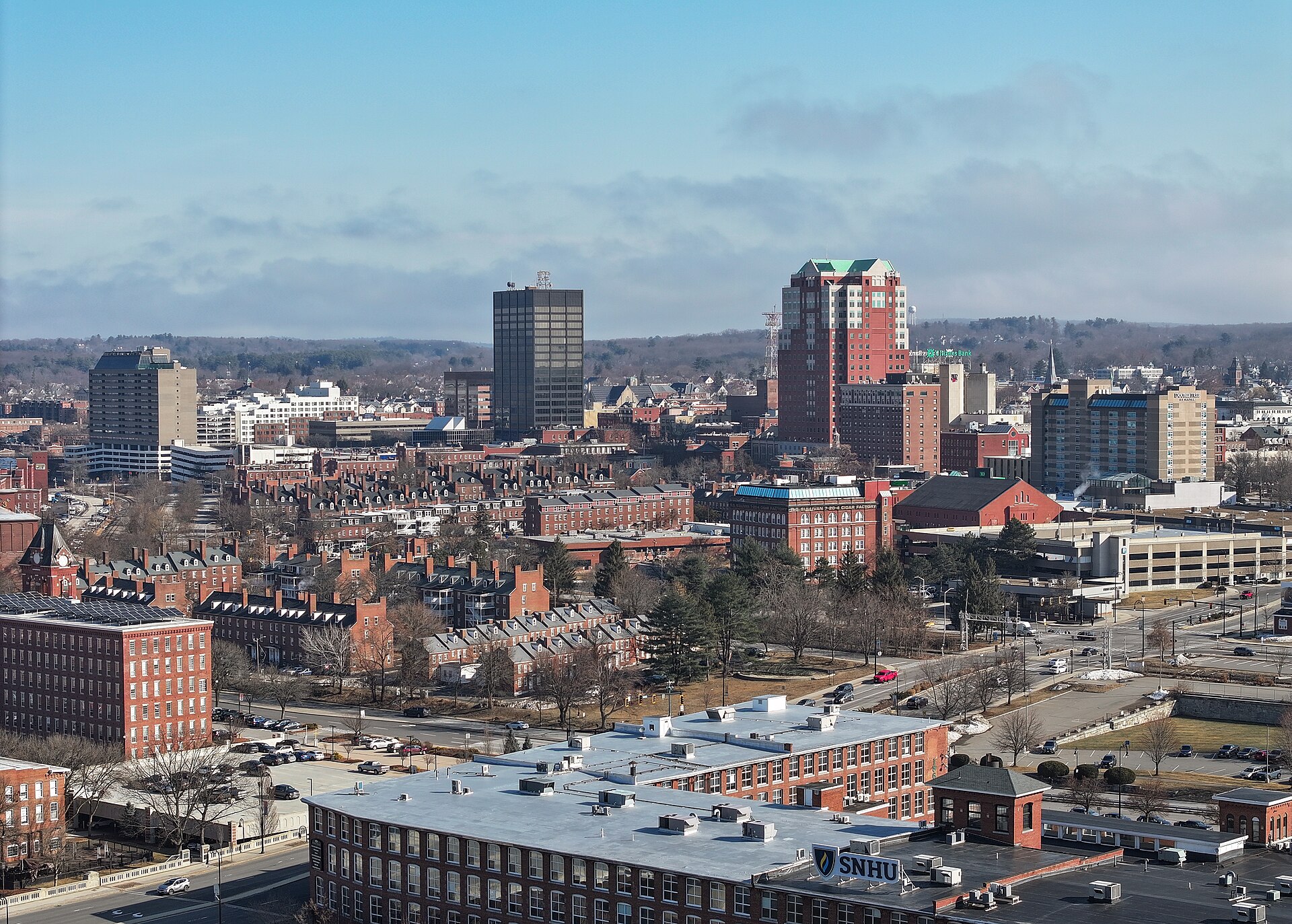 Downtown Manchester’s towers and brick rowhouses rising beyond the old Millyard mill buildings under a pale blue sky.