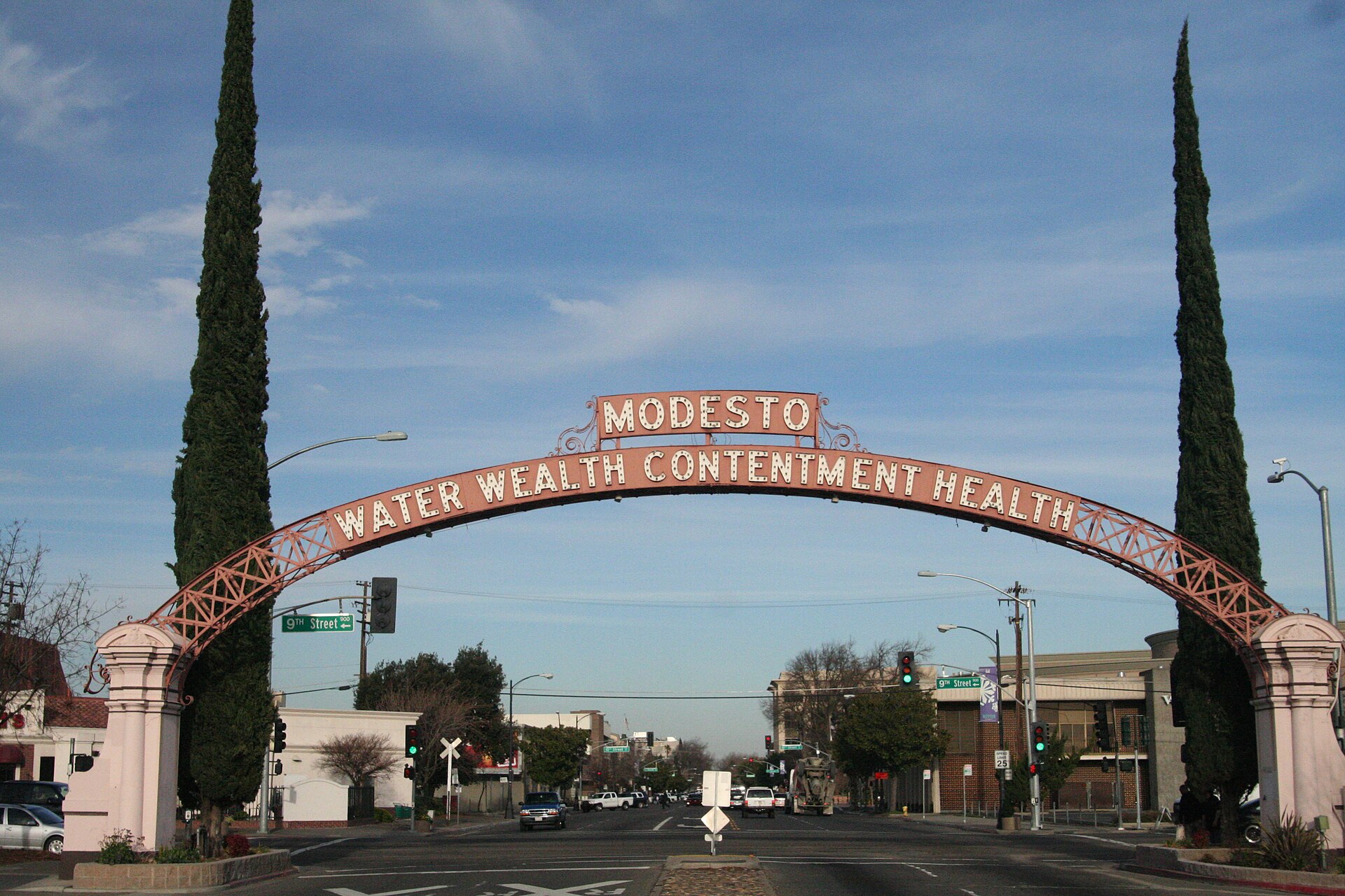 The Modesto Arch, reading “Water Wealth Contentment Health”, spanning a downtown street between tall cypress trees under a blue sky.
