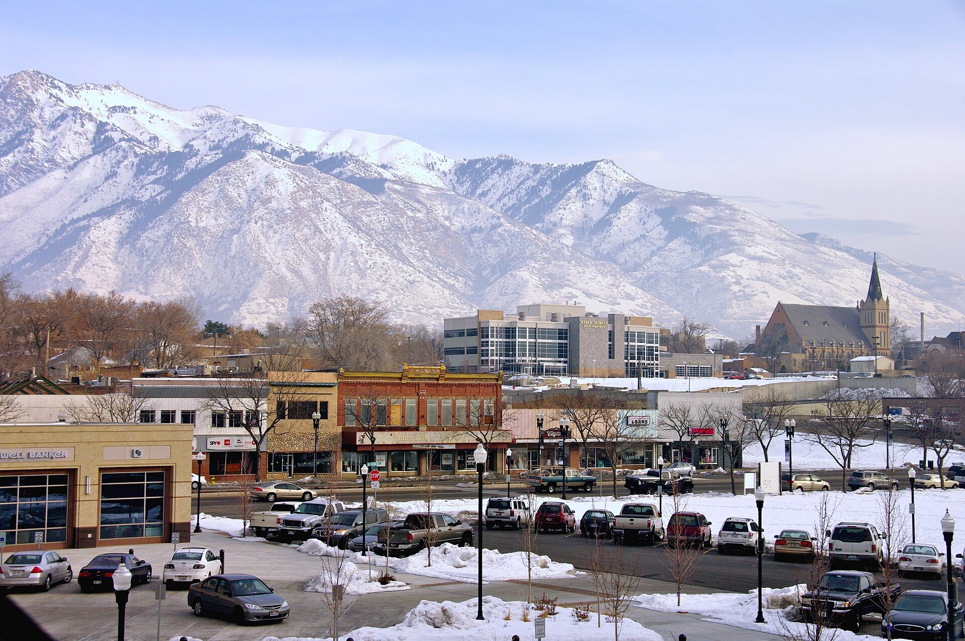 Downtown Ogden’s brick storefronts and a church spire beneath the snow-covered Wasatch Mountains on a clear winter day.