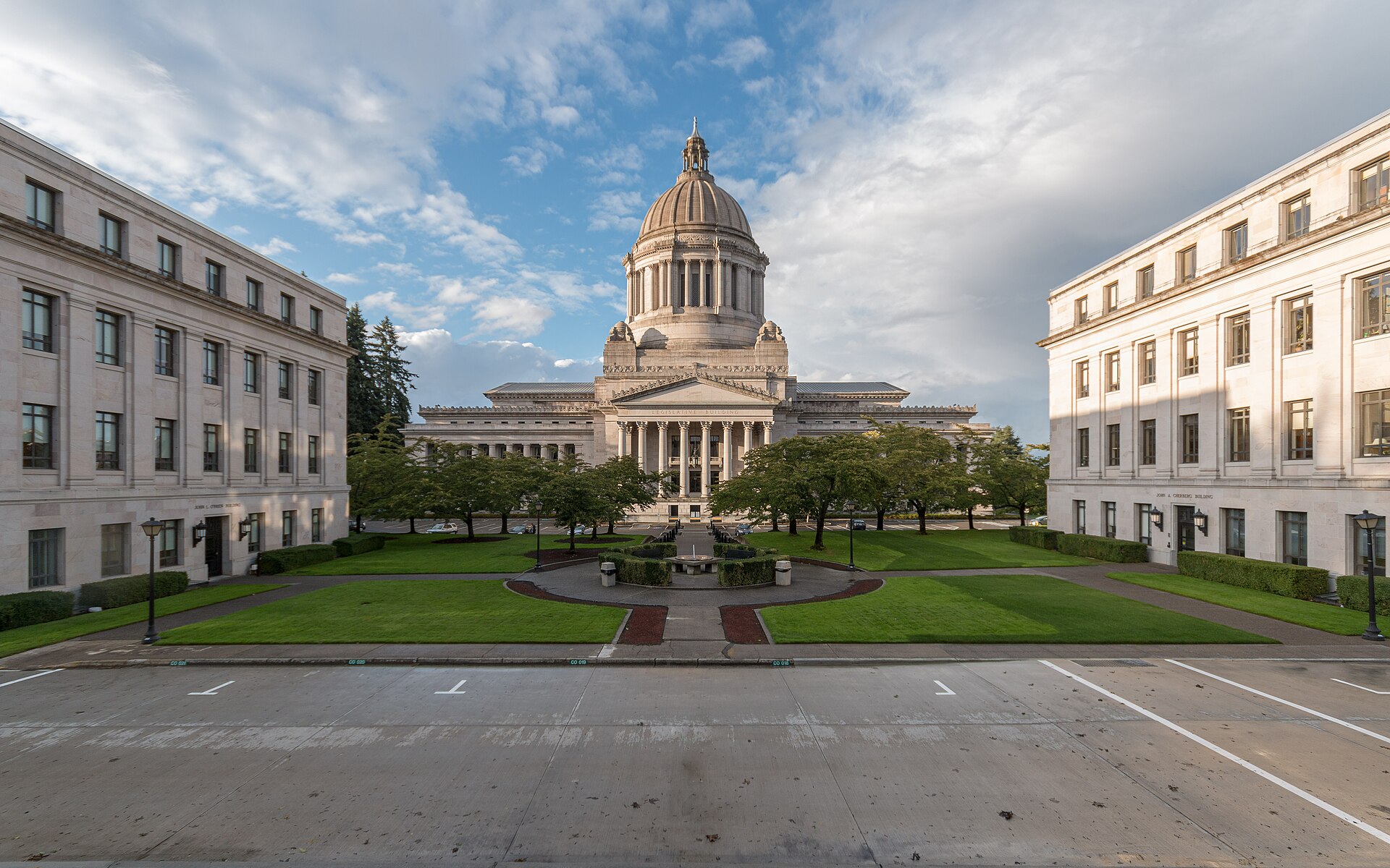 The domed Washington State Capitol in Olympia, framed by two stone office buildings and lawns under a partly cloudy sky.