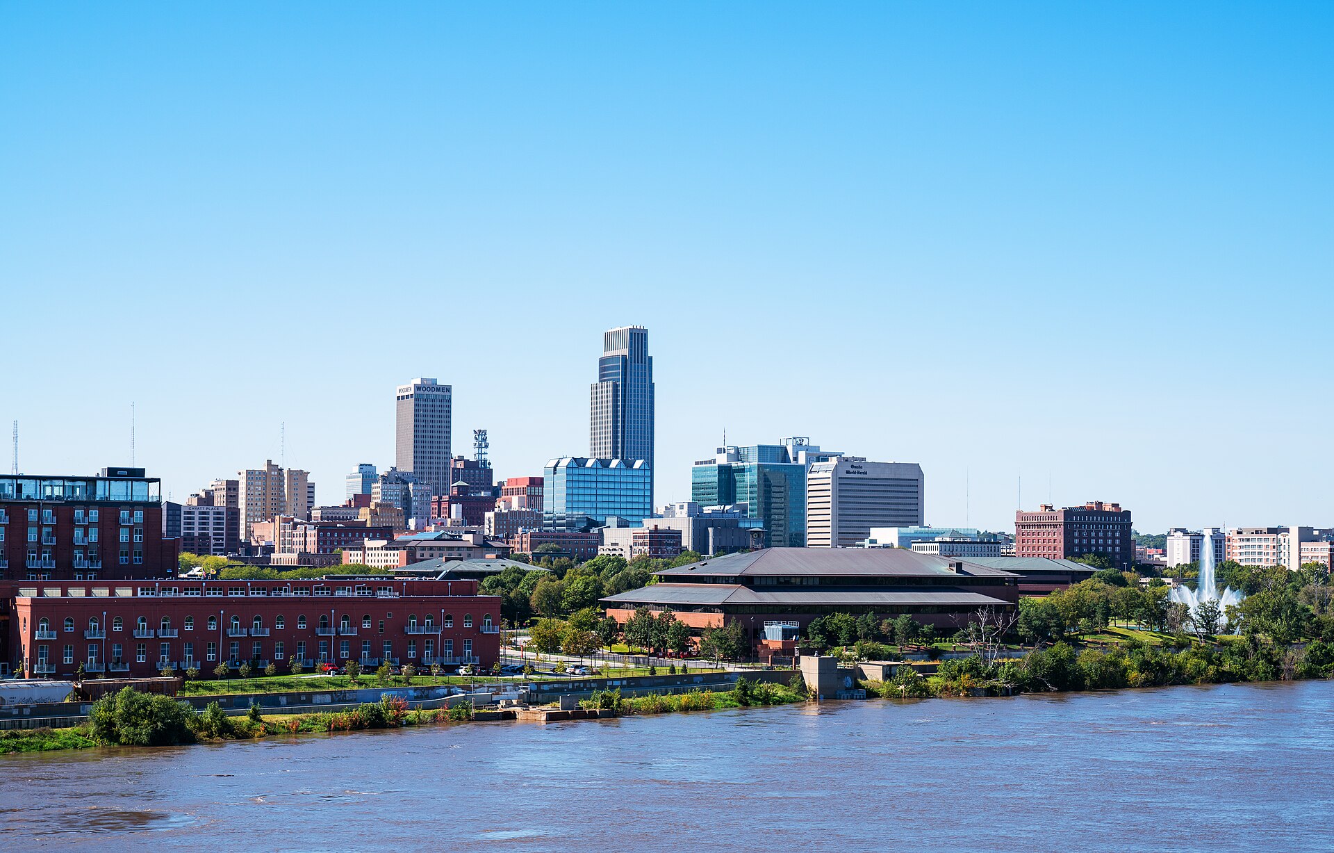 Downtown Omaha skyline rising above the Missouri River and green riverbank on a clear, sunny day.