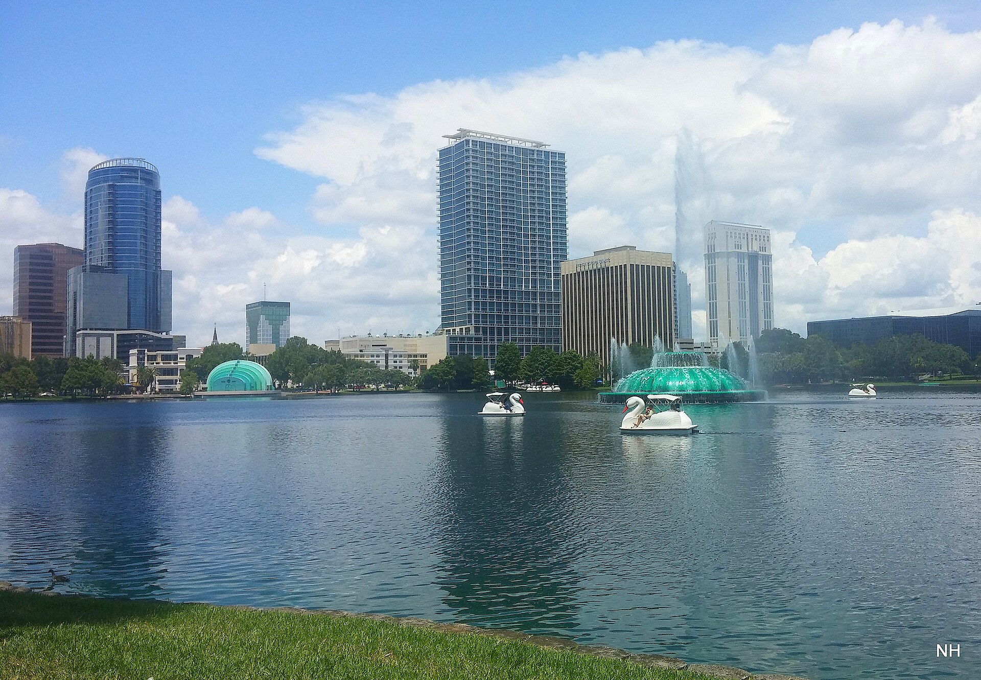 Swan boats and the fountain on Lake Eola, with downtown Orlando's towers rising behind under a bright sky.