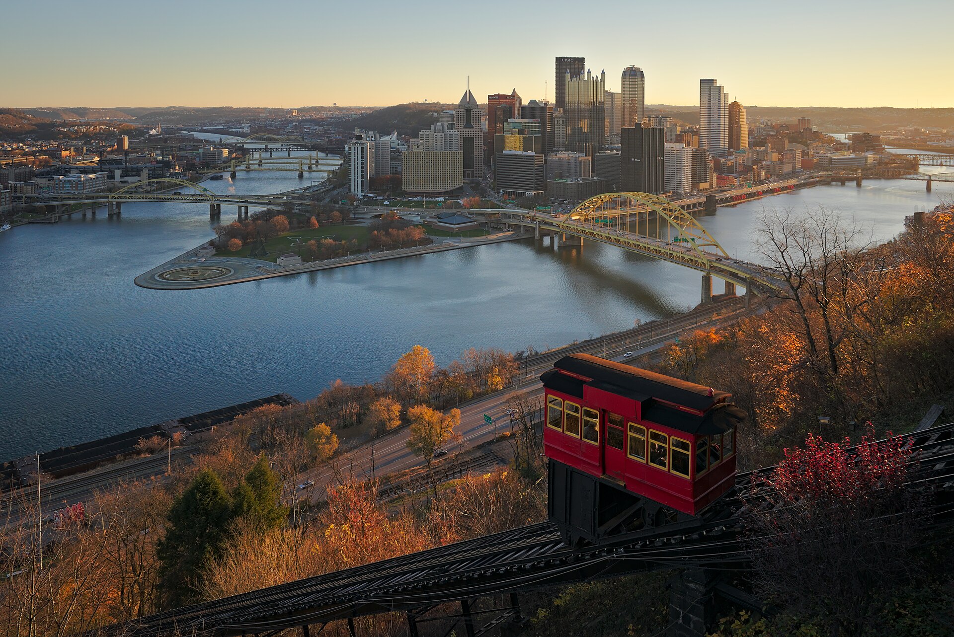 Downtown Pittsburgh's skyline and the Point where its rivers meet, seen from Mount Washington above a red incline car in morning sun.