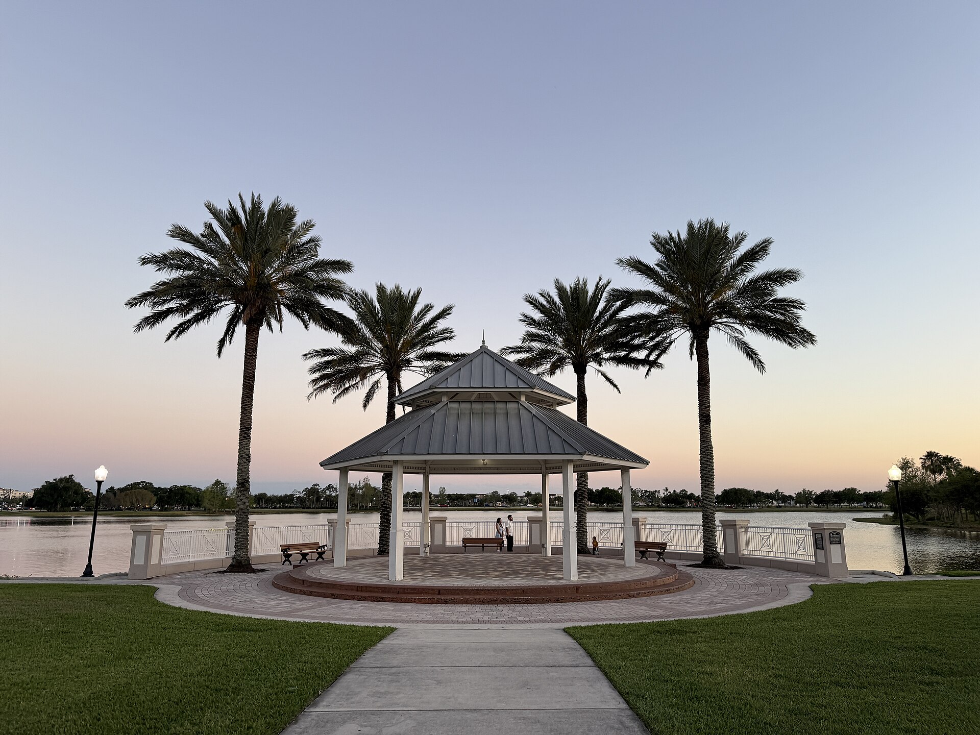 A white gazebo framed by four tall palm trees on the shore of Lake Tradition in Port St. Lucie at dusk.