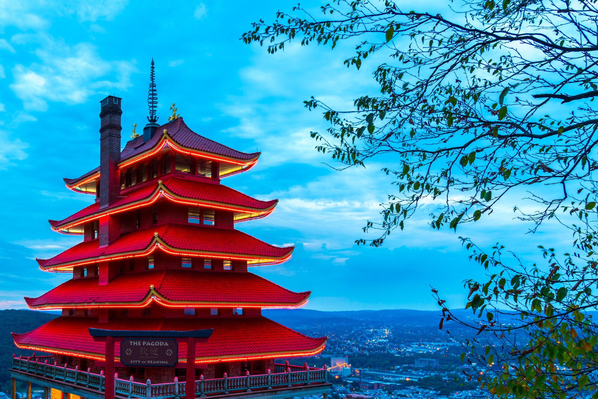 The red, tiered Pagoda on Mount Penn above Reading under a bright blue sky, framed by tree branches.