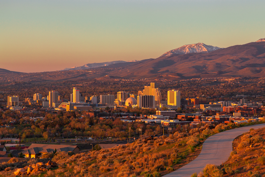 Downtown Reno's towers glowing in golden-hour light beneath snow-capped mountains, seen across a sagebrush hillside.