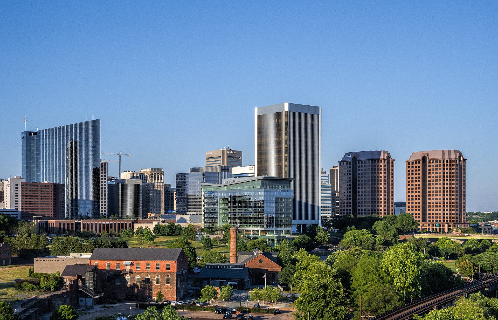 Downtown Richmond's office towers rise above trees and old brick buildings under a clear blue sky.