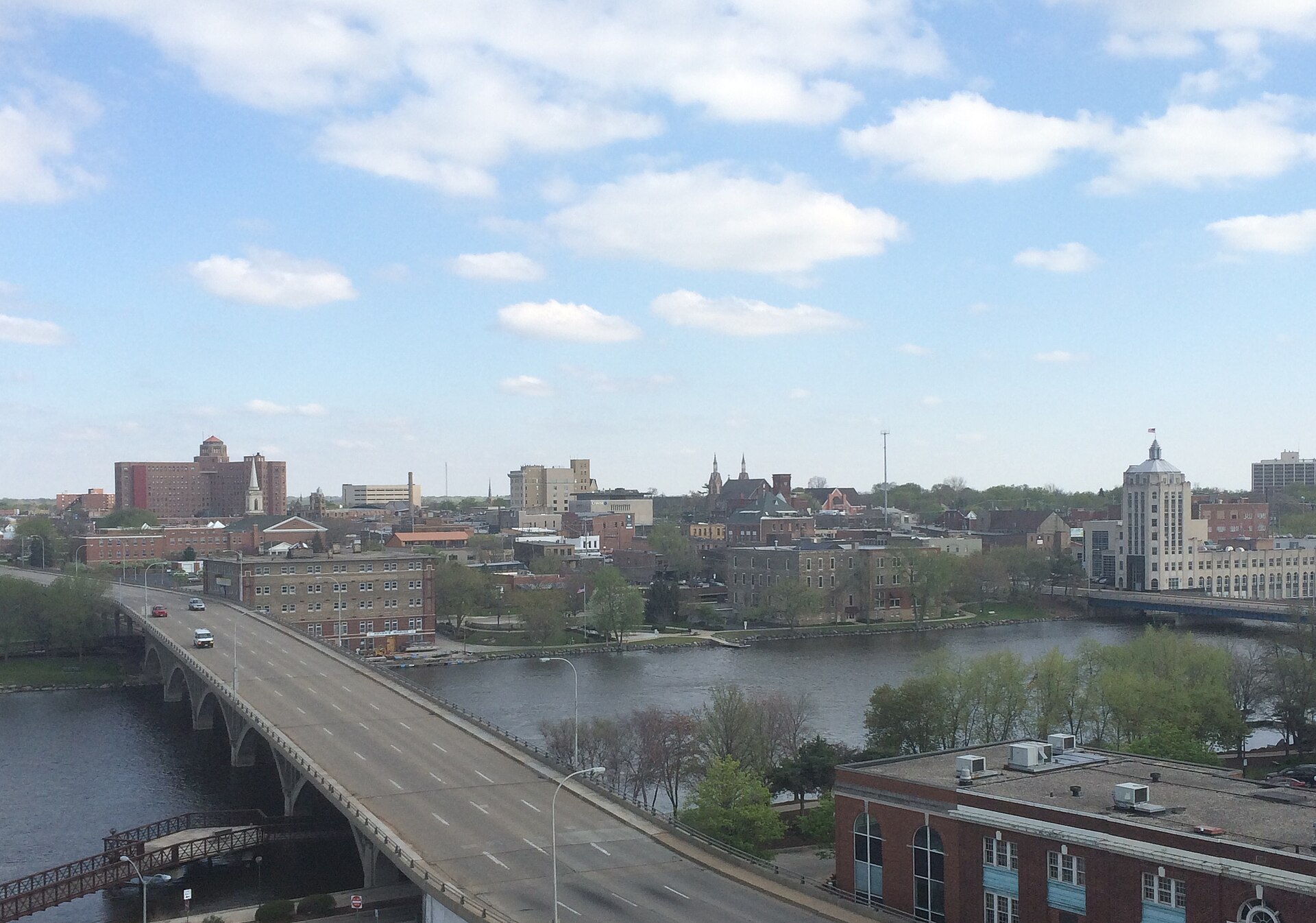 The Rock River winding past downtown Rockford's brick buildings and towers, with the Jefferson Street Bridge and a blue sky.
