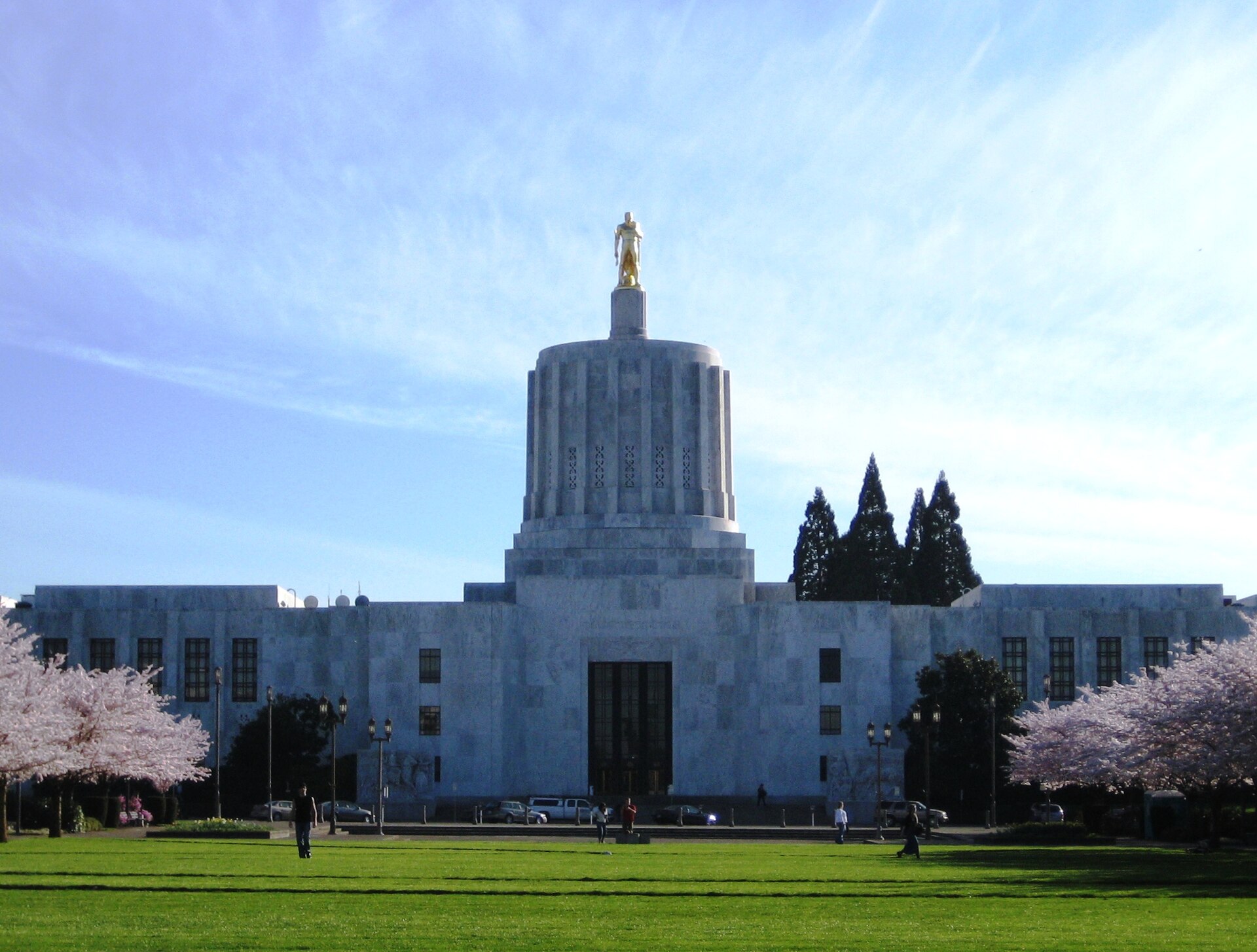 The white marble Oregon State Capitol with its gilded pioneer statue, framed by blossoming cherry trees on the lawn in Salem.
