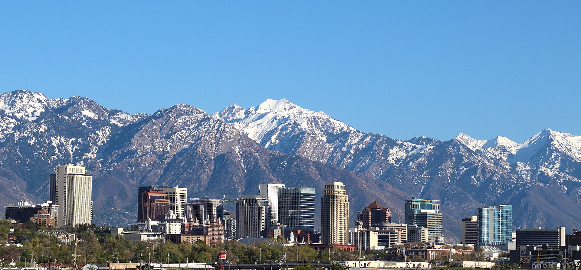 Downtown Salt Lake City's towers in front of the snow-capped Wasatch Mountains under a clear blue sky.
