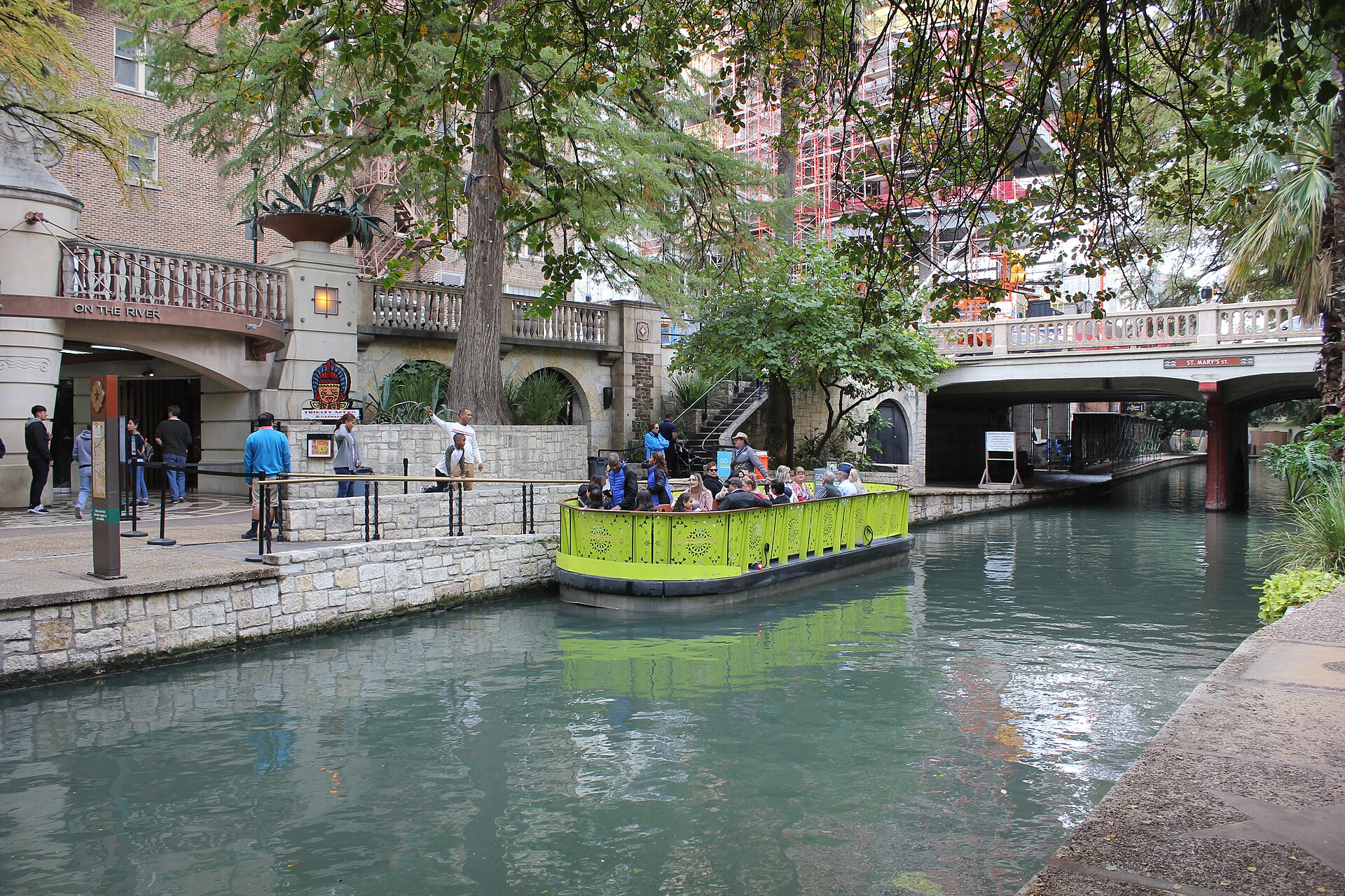 A tour barge gliding along the tree-shaded San Antonio River Walk past stone walls, stairs and an arched bridge.