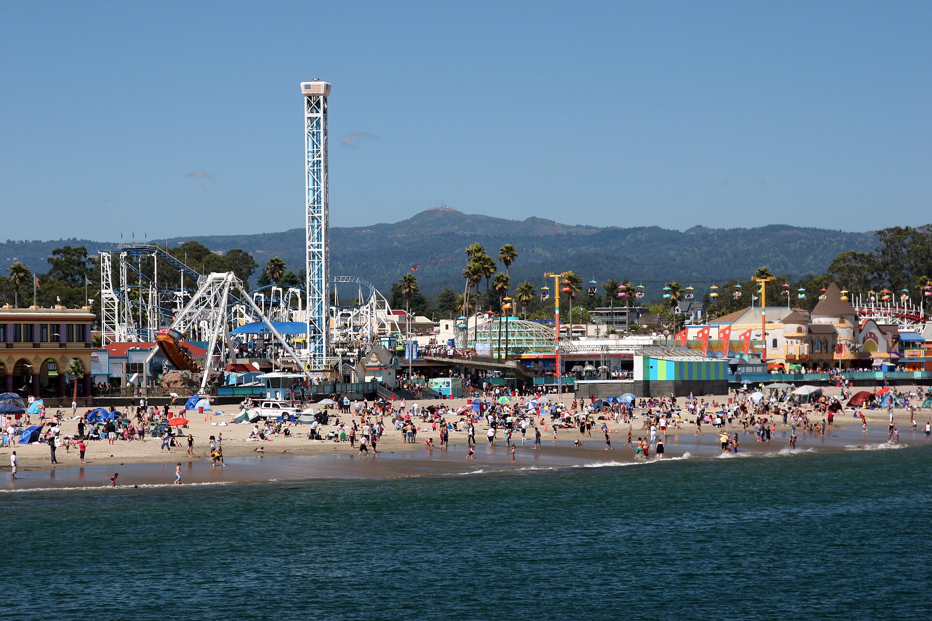 Rides of the Santa Cruz Beach Boardwalk rise above a busy beach on Monterey Bay, with wooded hills behind, on a sunny day.