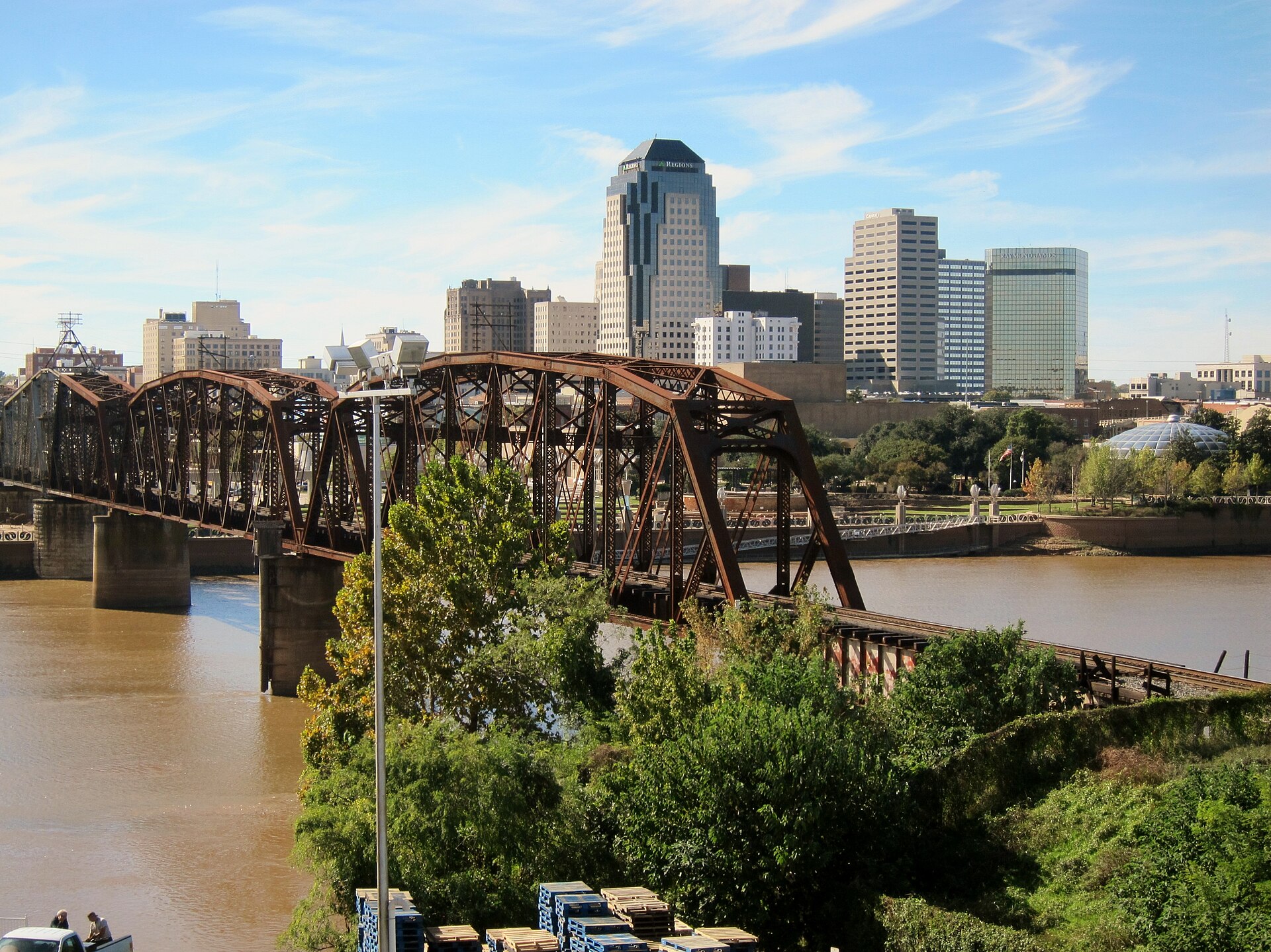 An old steel railroad bridge crosses the Red River in front of downtown Shreveport's skyline on a sunny day.