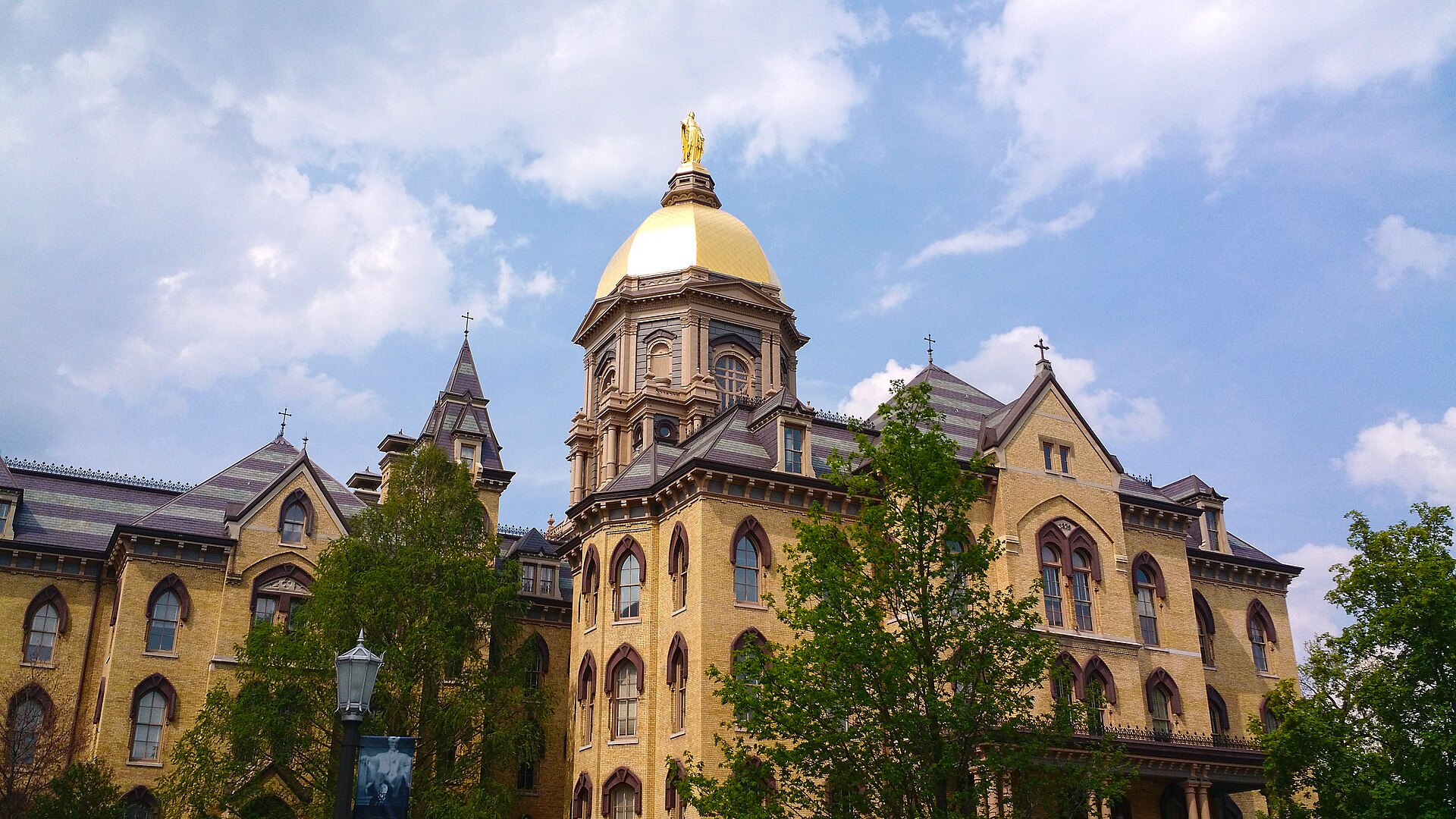 The gold-domed Main Building of the University of Notre Dame near South Bend, framed by trees under a blue sky.