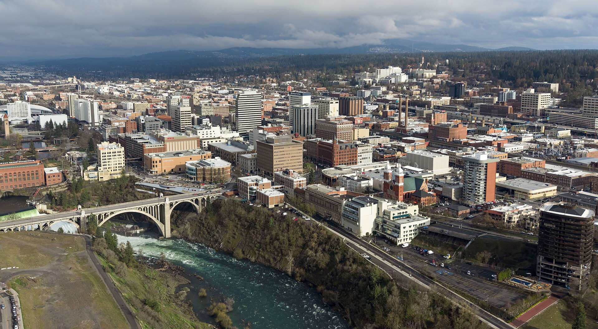 Downtown Spokane and the arched Monroe Street Bridge above the rushing Spokane River, seen from above on a cloudy day.