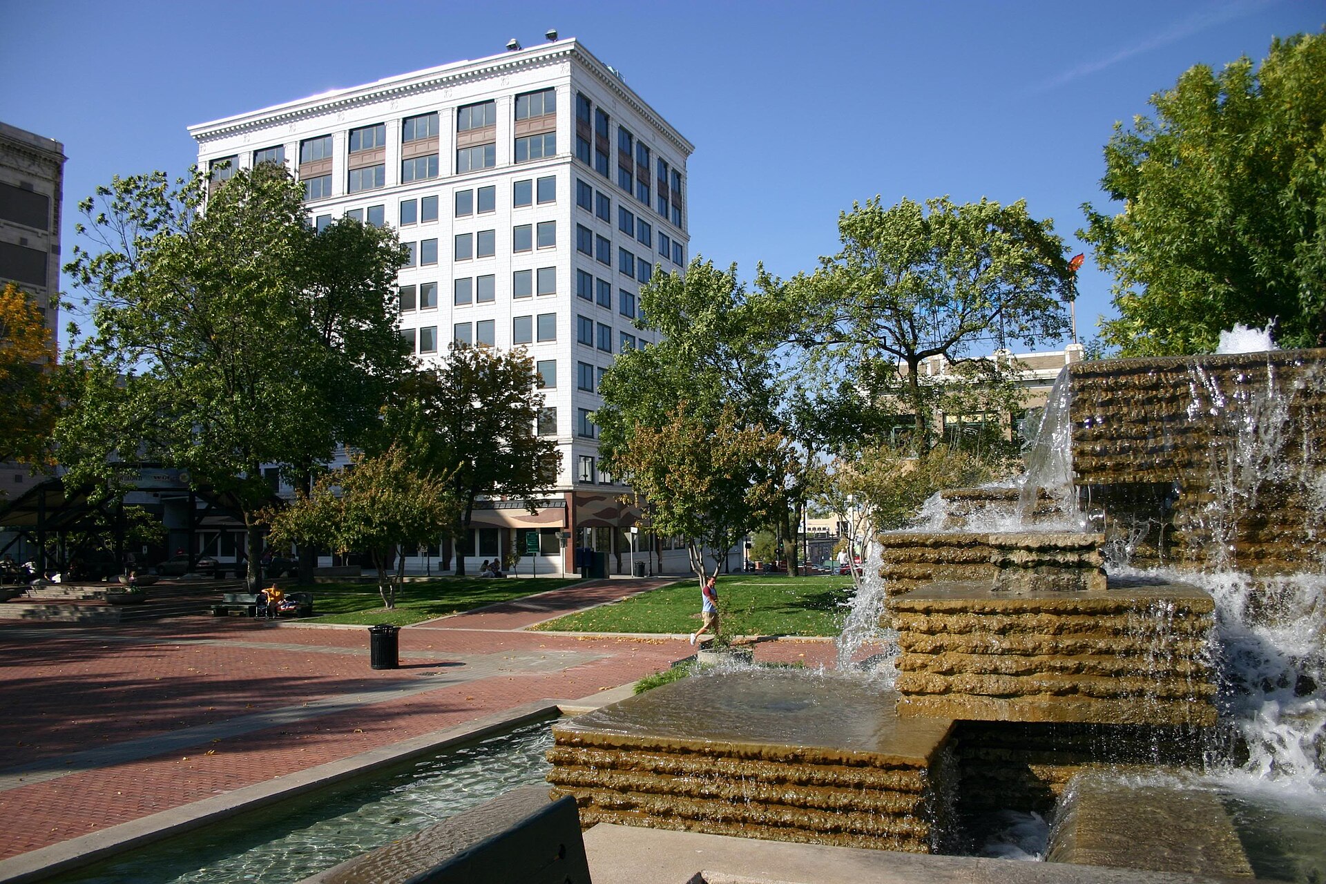 The stepped fountain, brick plaza and trees of Park Central Square, with a white early-1900s office tower, in downtown Springfield.