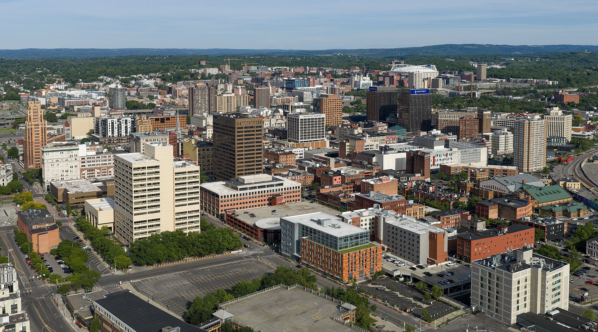 Downtown Syracuse office towers and brick buildings with green hills beyond, seen from above on a sunny day.