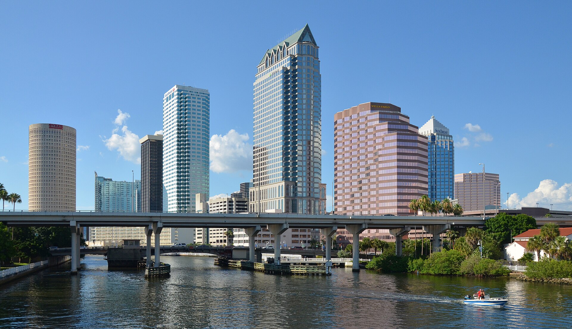Downtown Tampa's skyscrapers rise behind a highway bridge over the Hillsborough River on a sunny day.