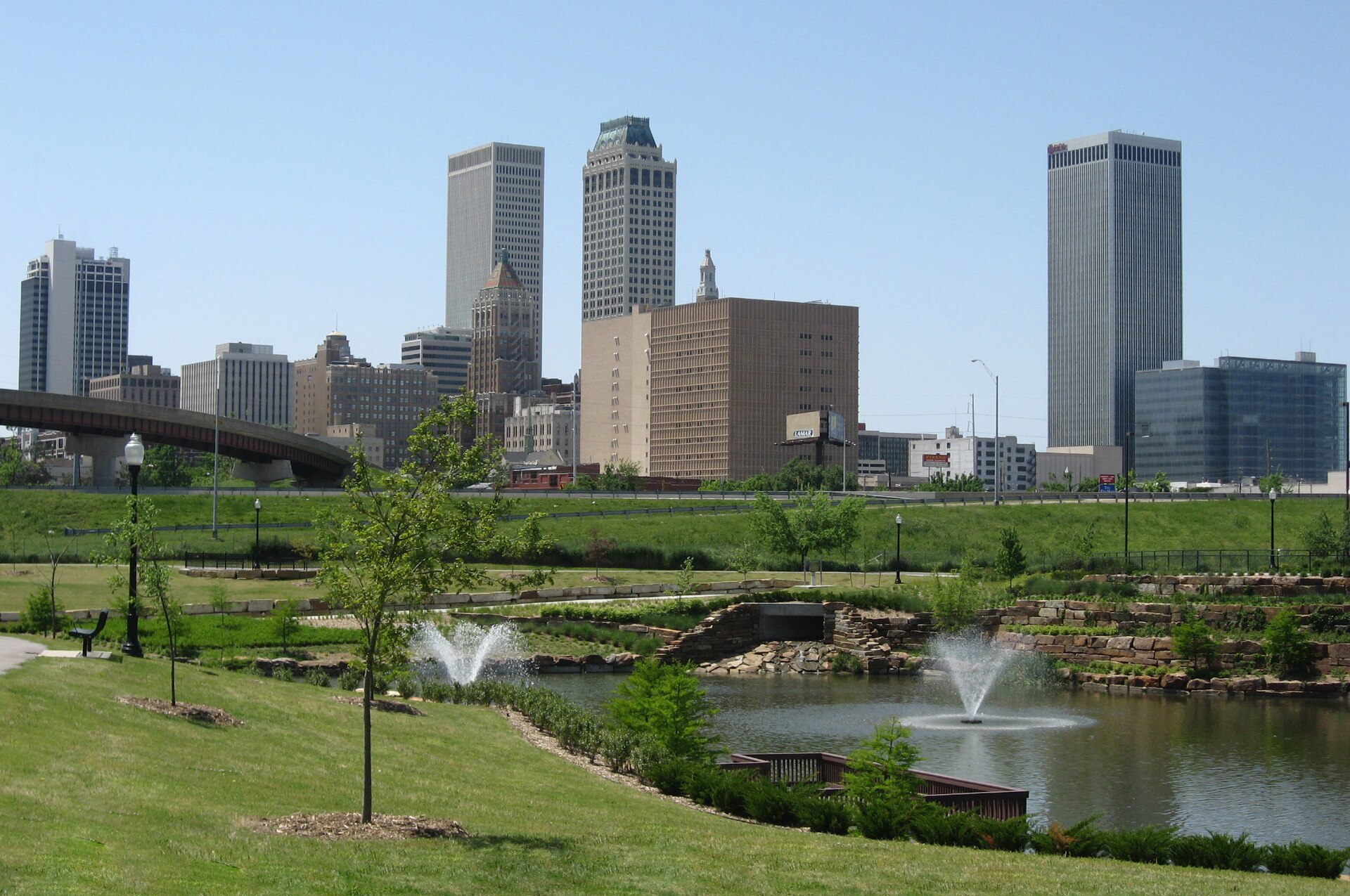 Downtown Tulsa’s towers rising beyond a green park with a pond and fountains on a clear spring day.