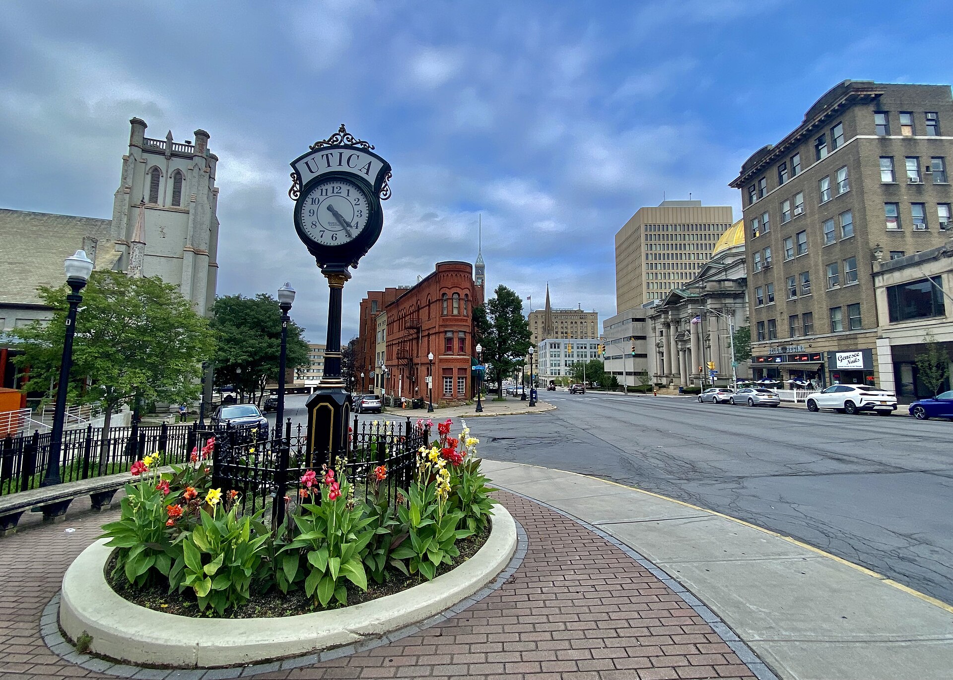 A vintage street clock reading Utica above a flower bed on Genesee Street, with historic brick buildings and a church tower.