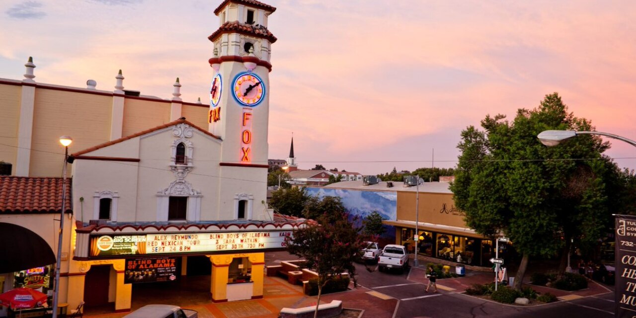 The Visalia Fox Theatre's clock tower and glowing marquee on a downtown Visalia corner under a pink dusk sky.